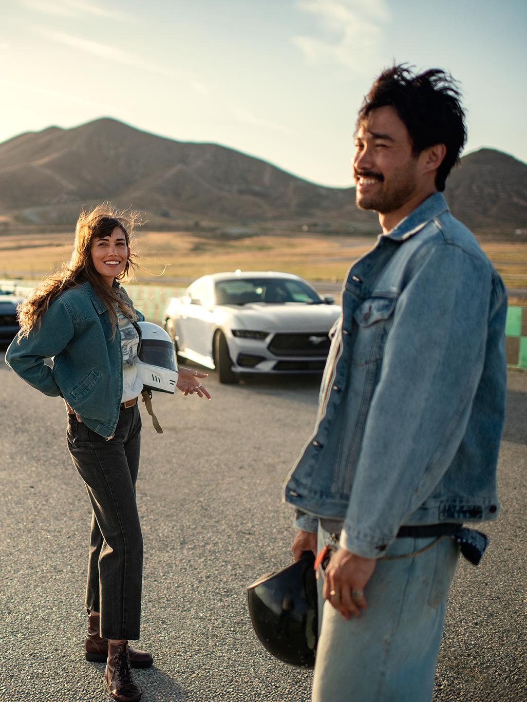 Two people stand near a Ford Mustang parked on a gravel road.