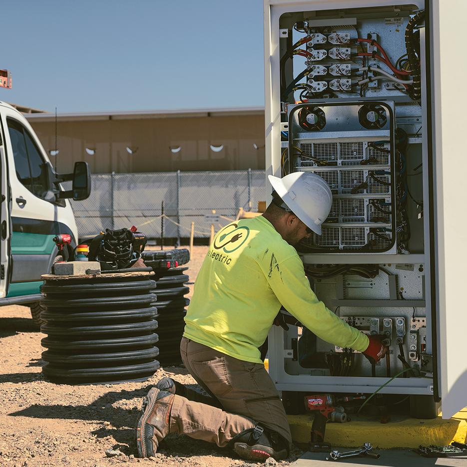A male construction worker works on an electrical system at a construction site, with a Ford Transit partially visible on the right. 