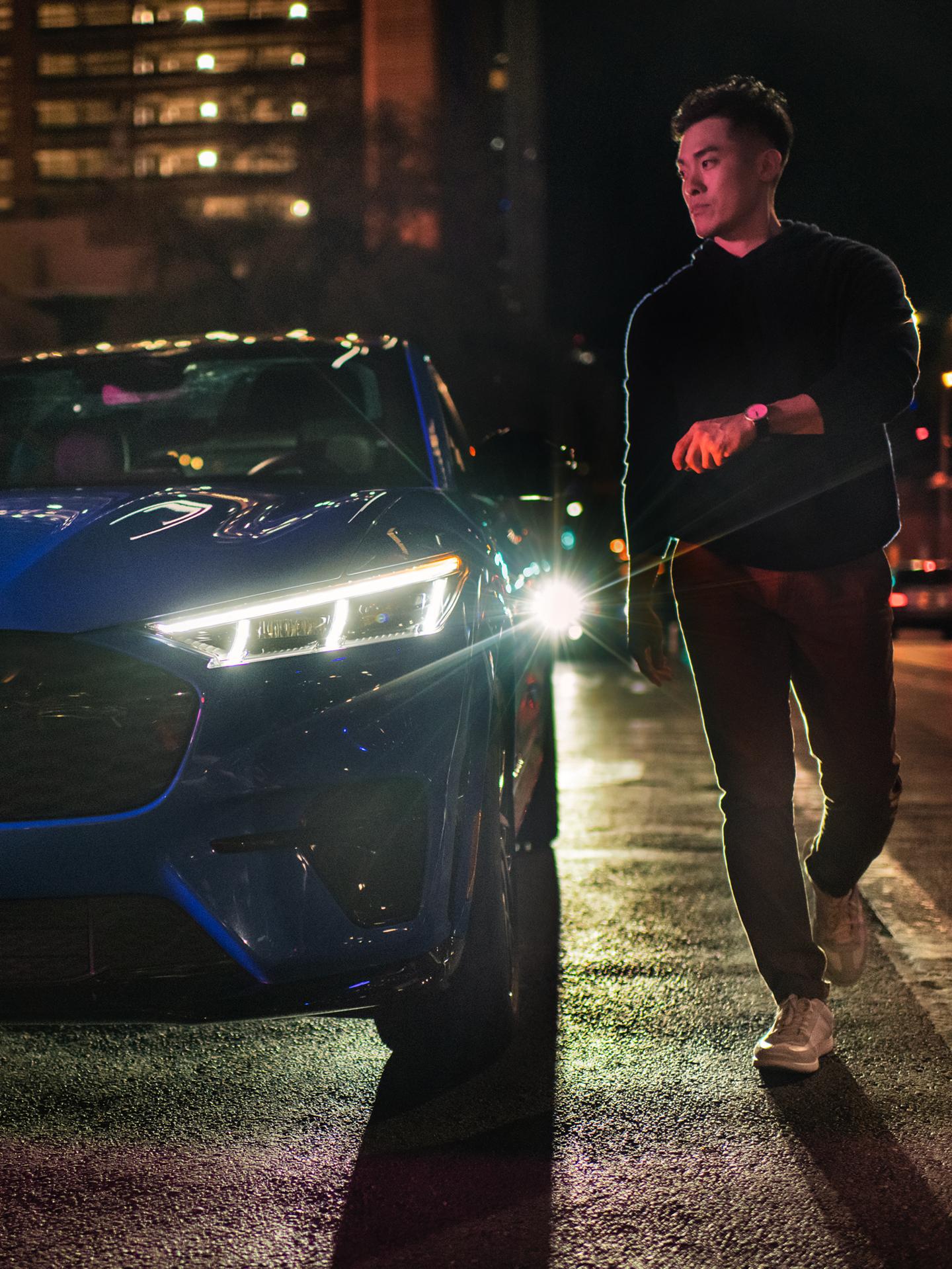 Person walking toward a Mustang Mach-E parked on an urban street at night.