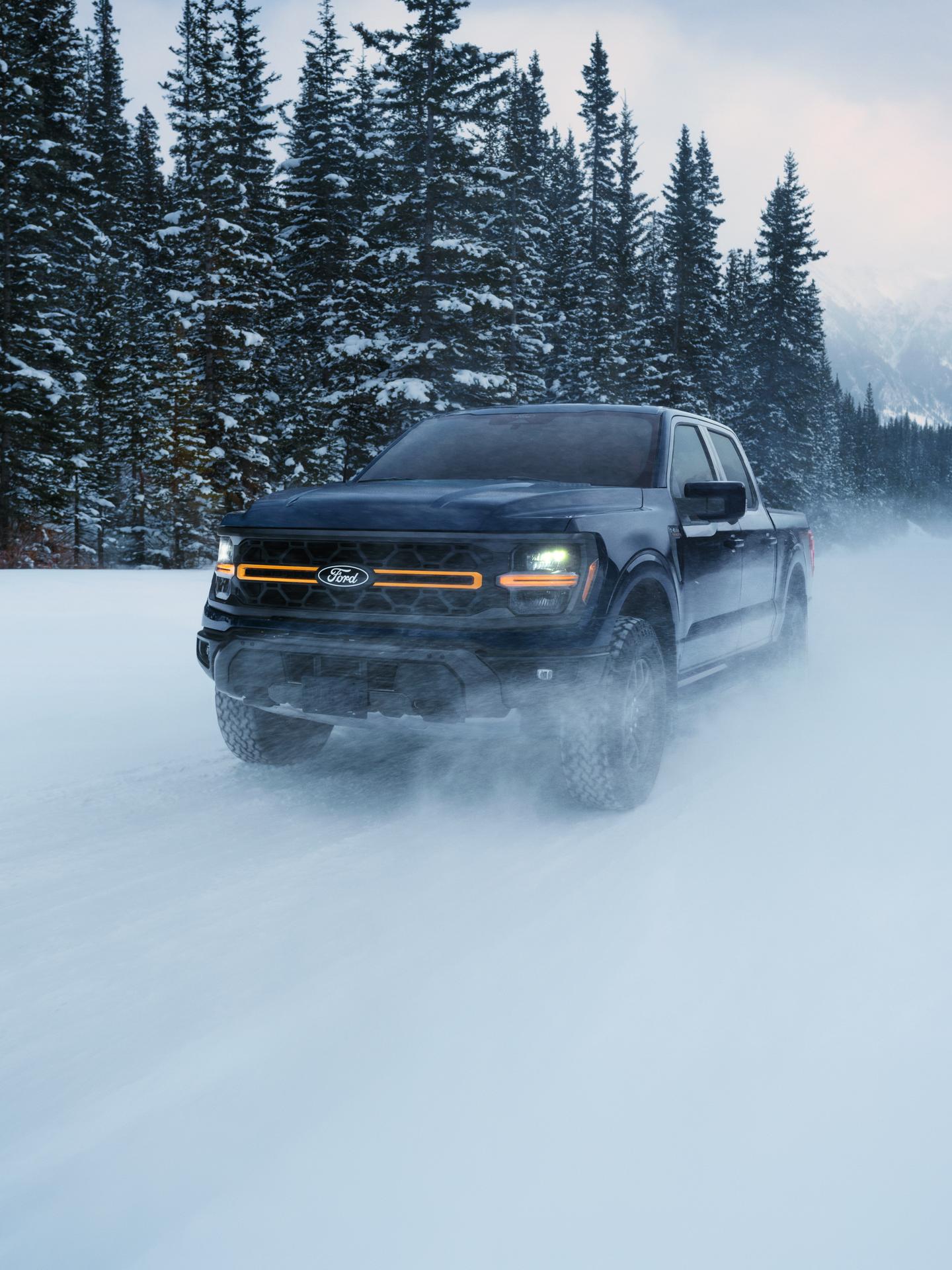 A blue Ford F-150 driving on snowy trail with pine trees in the background.