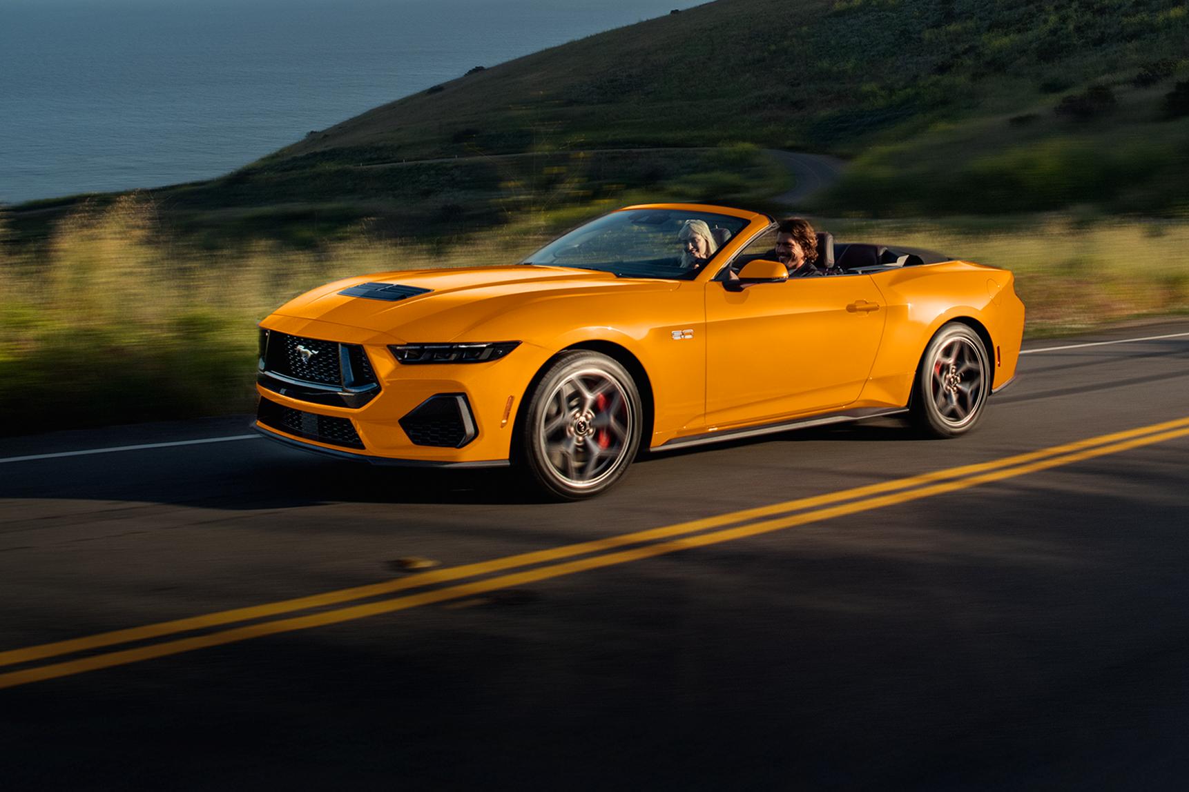 Two people in a 2026 Ford Mustang® GT Convertible in Orange Fury Metallic Tri-coat being driven on a coastal highway