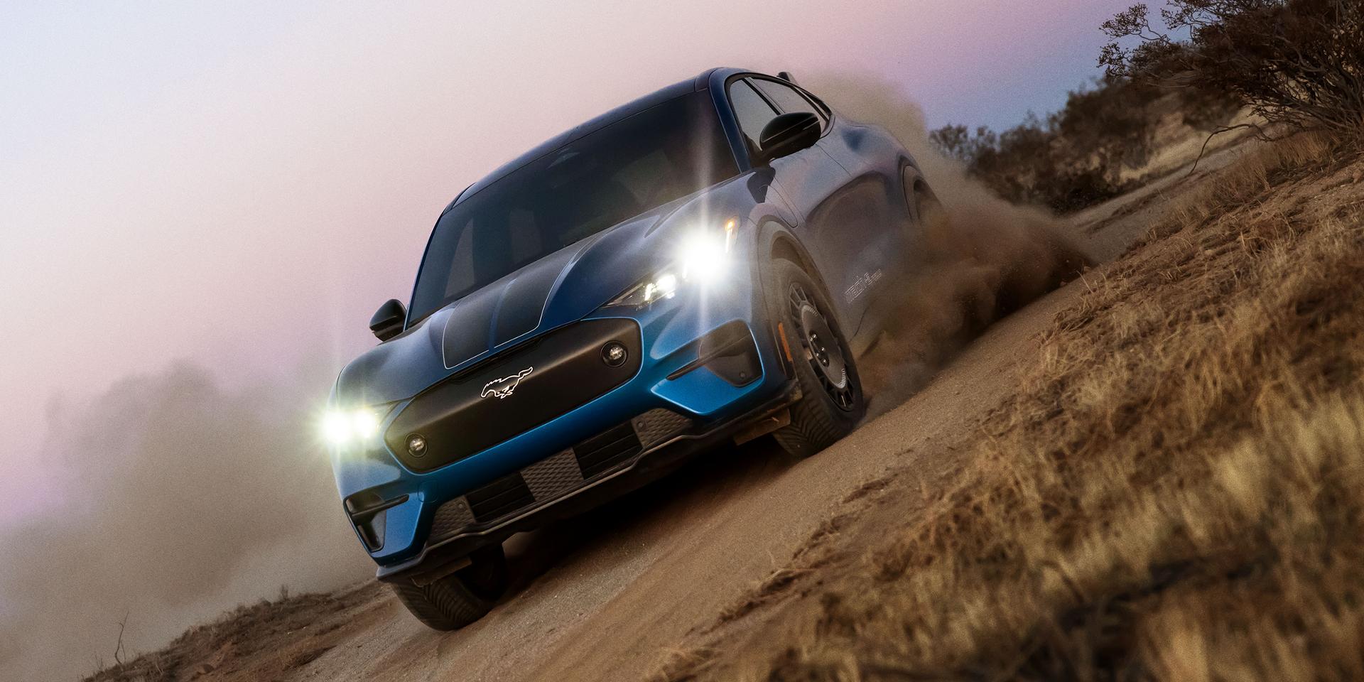 Three-quarter front view of a blue Ford Mustang Mach-E electric SUV driving on a dirt road at an angle.