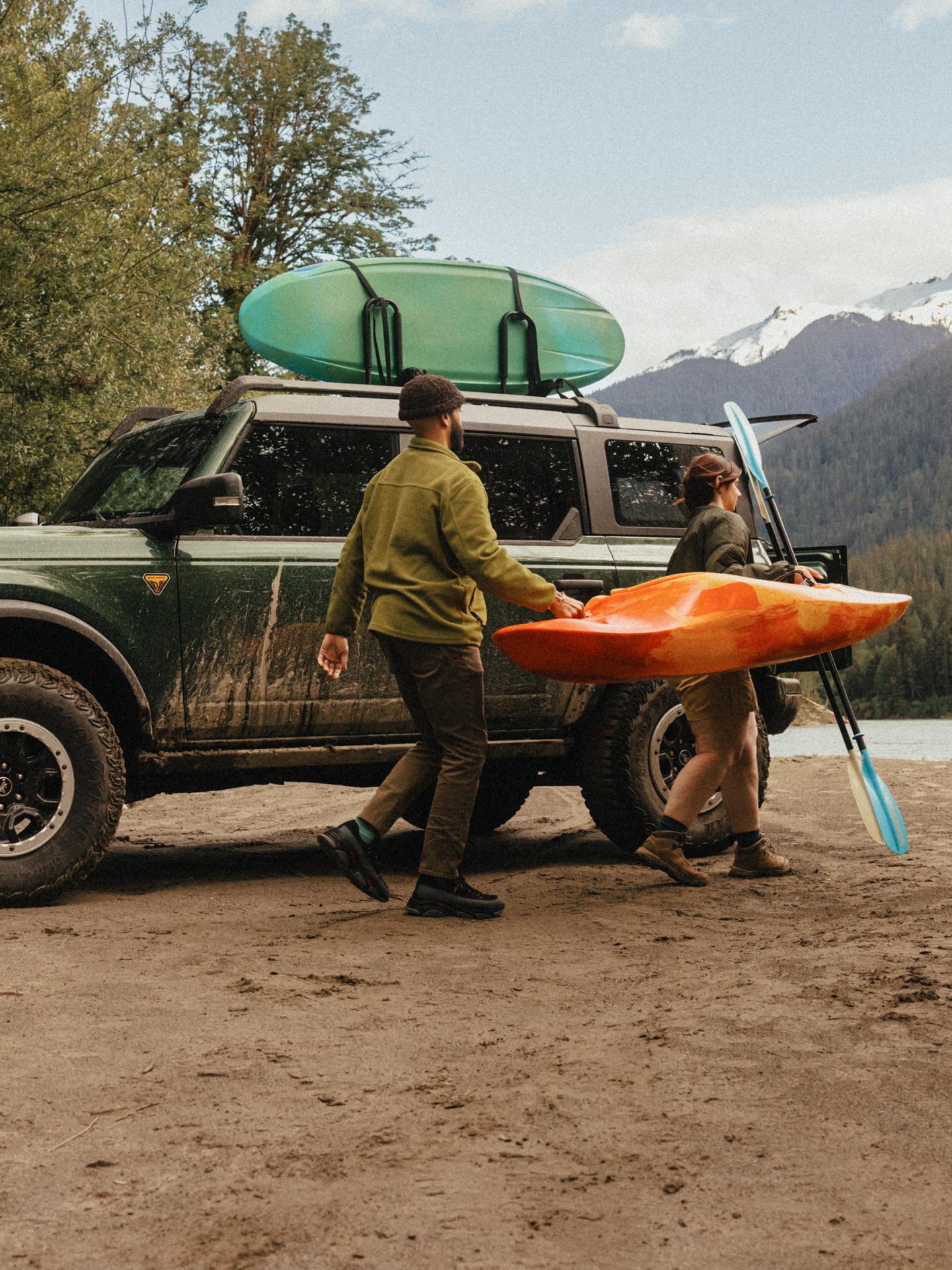 Two people carry a kayak past a Ford Bronco parked by a lake.