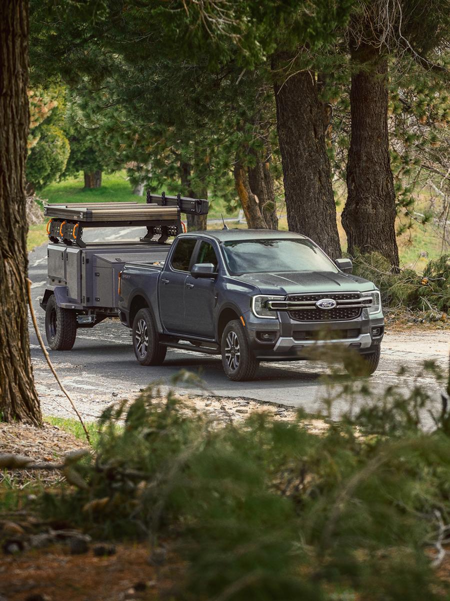 A three-quarter front shot of a dark gray Ford Ranger truck towing an off-road camper trailer along a wooded road.