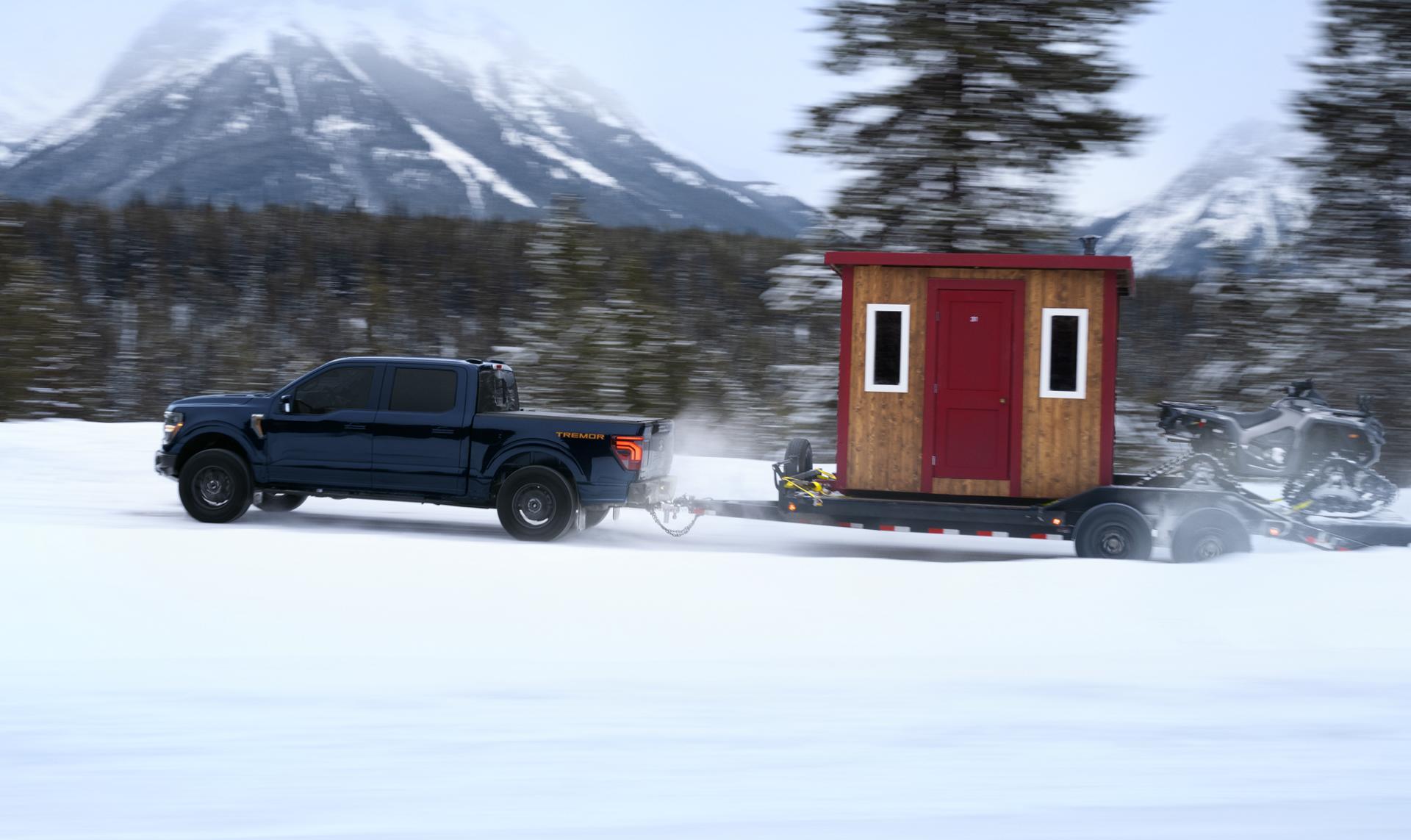 A dark blue Ford F-150 tows a fishing cabin on a snow-covered mountain trail.