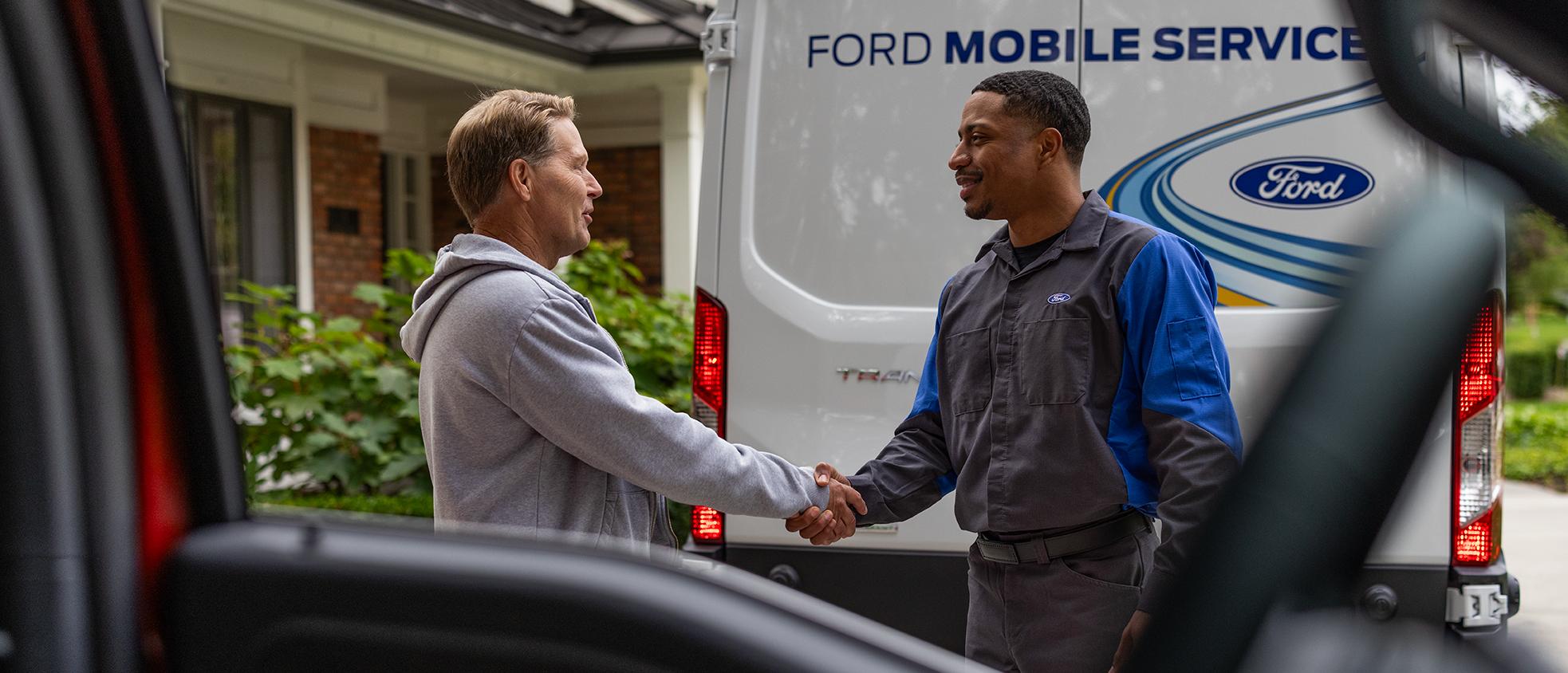 A Ford owner shakes hands with a Ford Mobile Service technician, framed through the window of a Ford vehicle.