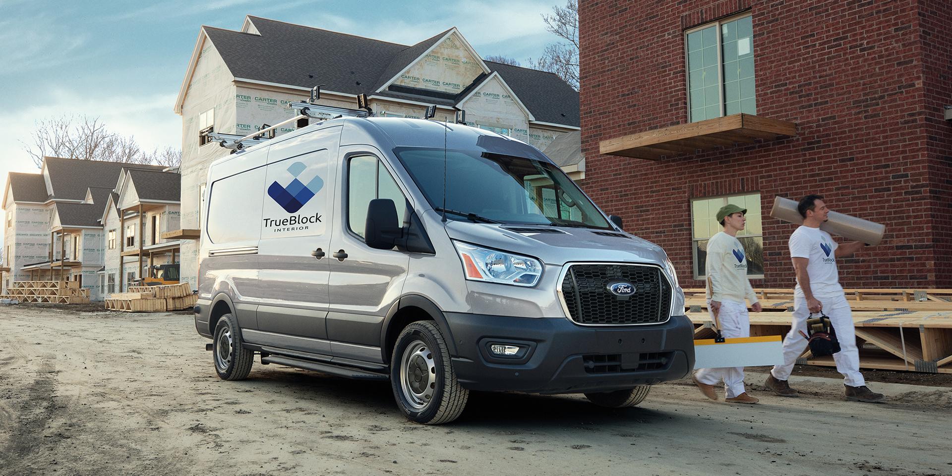 A three-quarter side view of a grey 2025 Ford Transit with two male construction workers walking in front of it at a job site.   