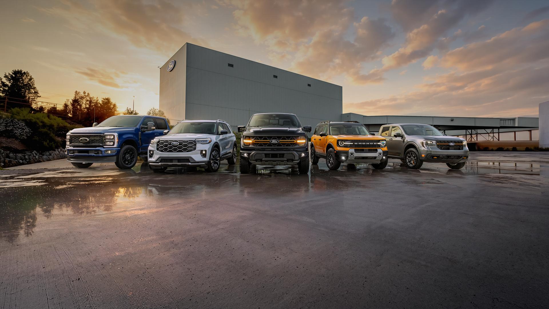 A range of Ford vehicles are parked in front of a Ford plant on rain-soaked asphalt under a cloudy sky at sundown.