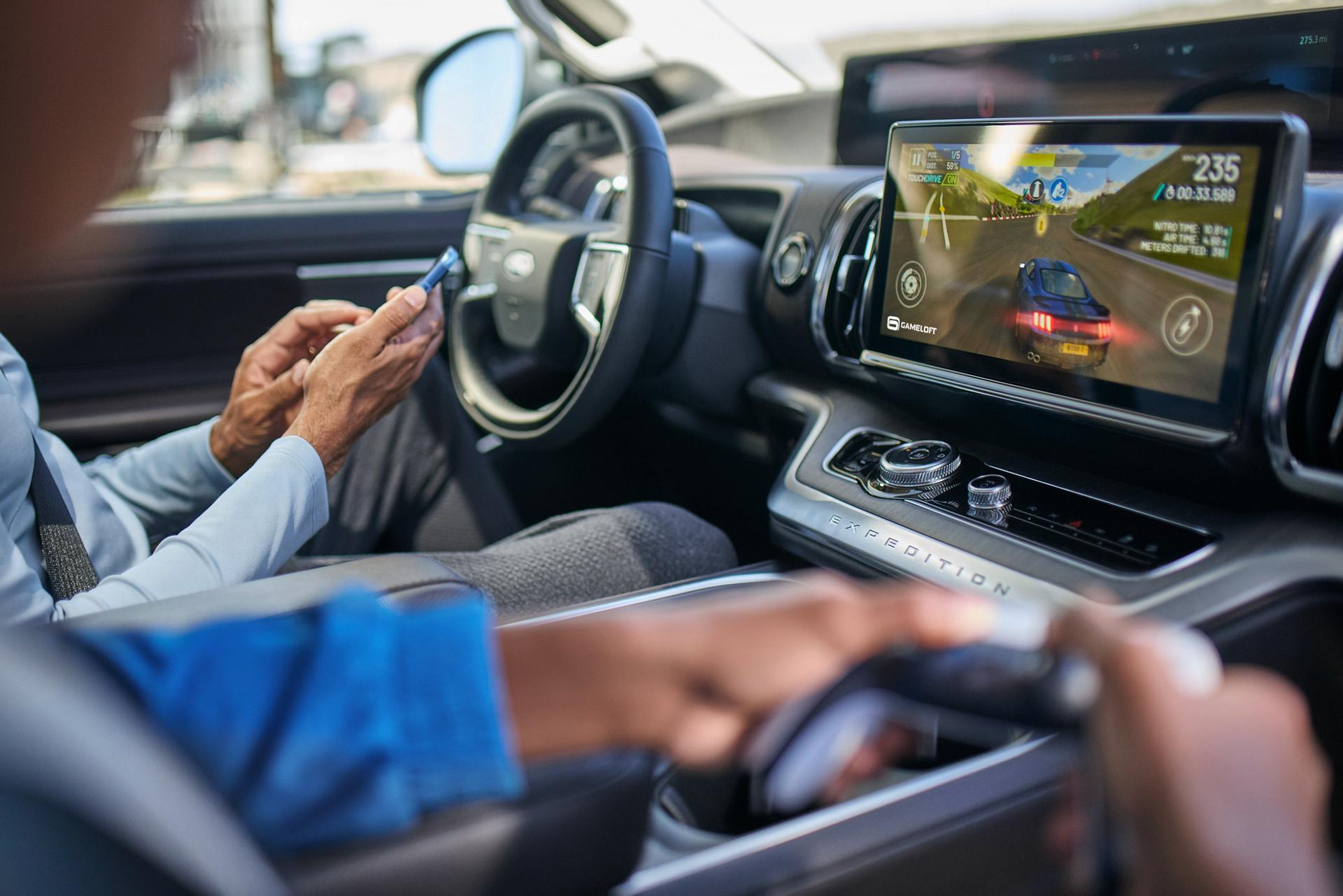 Driver and passenger in a parked Ford vehicle while the passenger plays a video game on the centre screen.