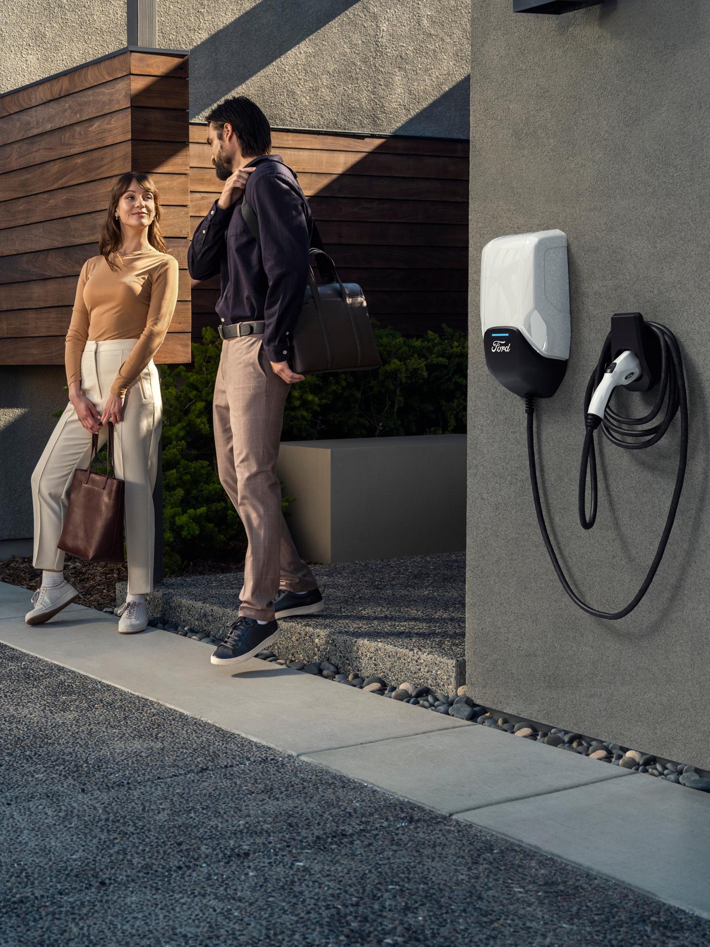 A couple chat in front of a home with a Ford Charge Station Pro mounted on a nearby wall.