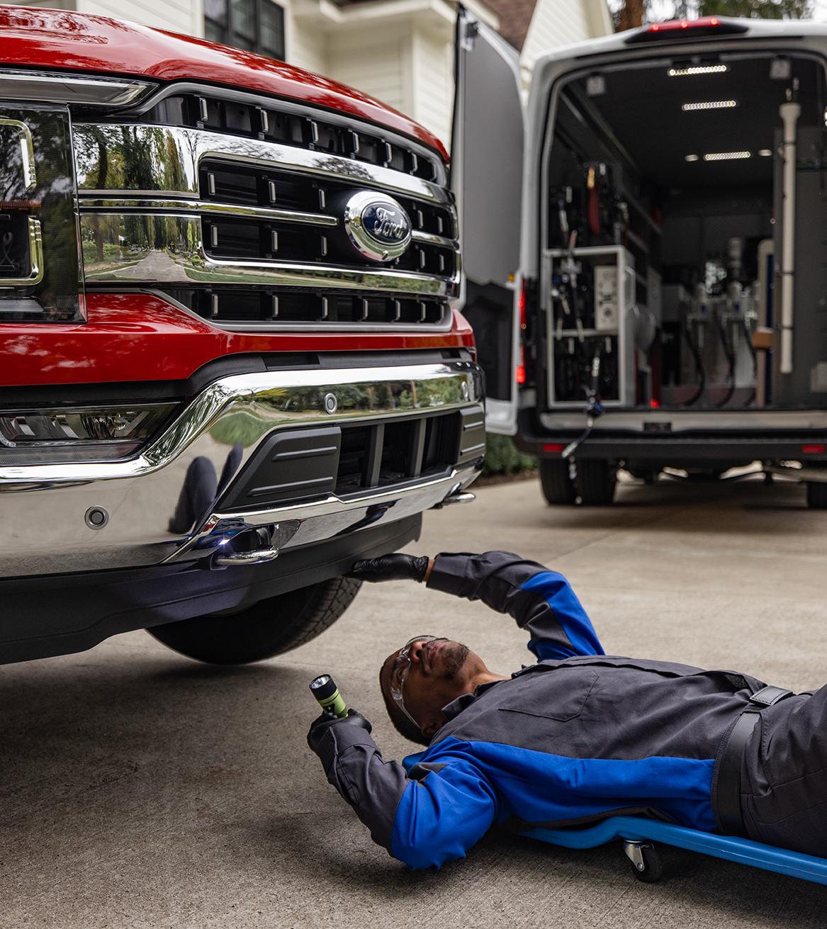A Ford Service technician on a mechanic’s creeper inspecting the underside of a F-150 in a customer’s driveway.