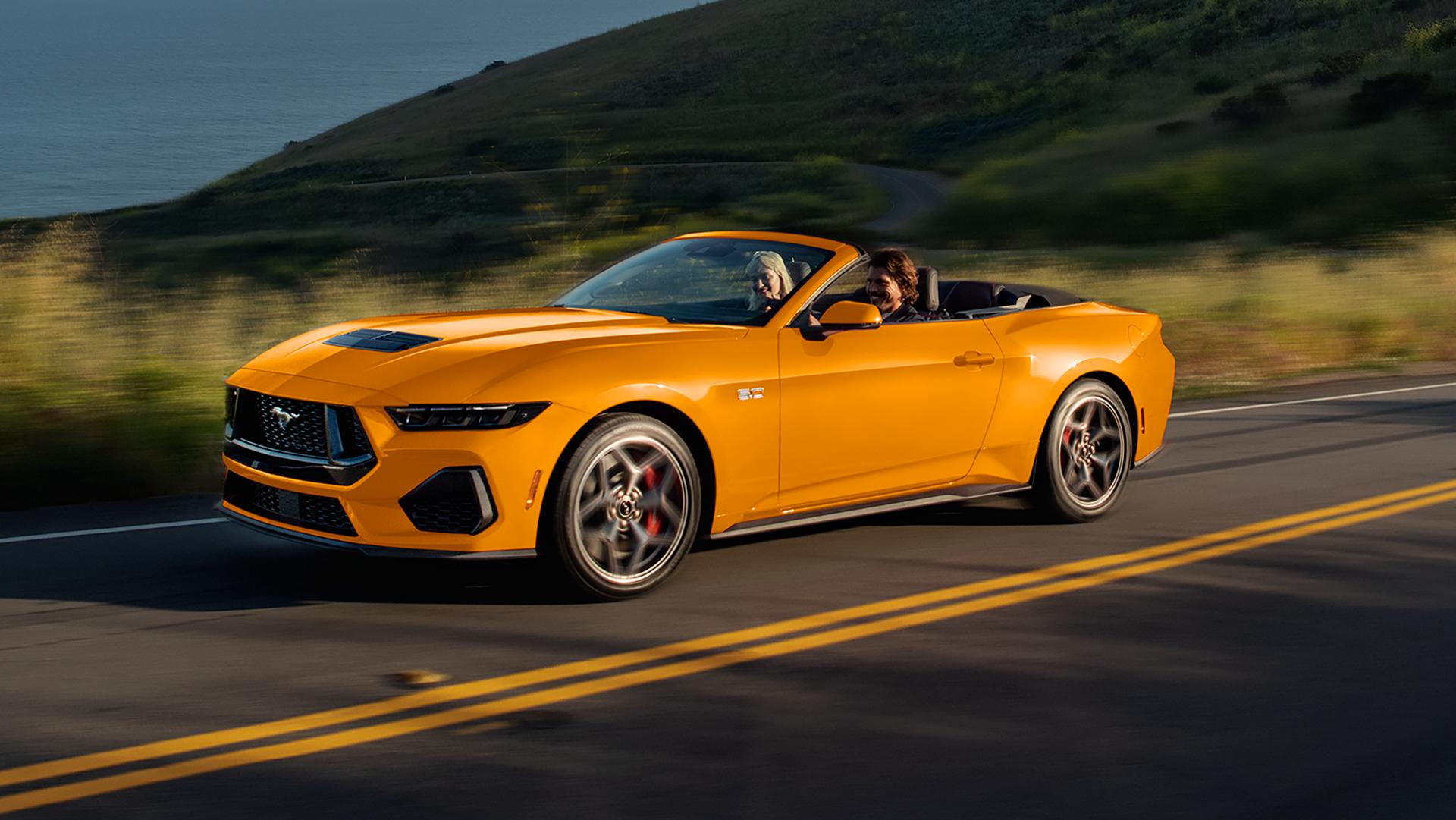 Two people in a 2026 Ford Mustang® GT Convertible in Orange Fury Metallic Tri-coat being driven on a coastal highway
