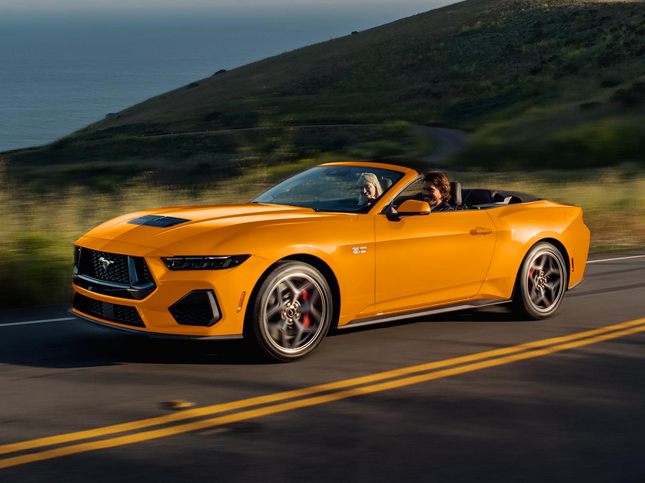 Two people in a 2026 Ford Mustang® GT Convertible in Orange Fury Metallic Tri-coat being driven on a coastal highway