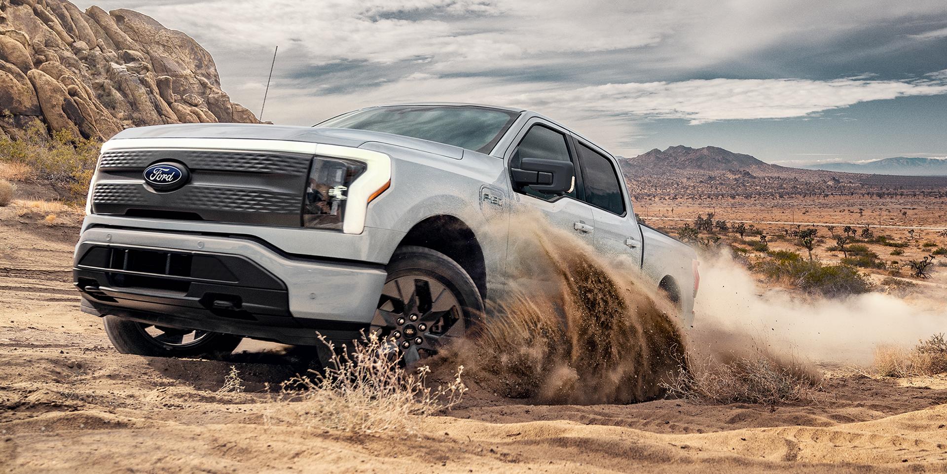 A three-quarter side view of a white F-150 Lightning kicking up dust as it charges through desert-like terrain. 
