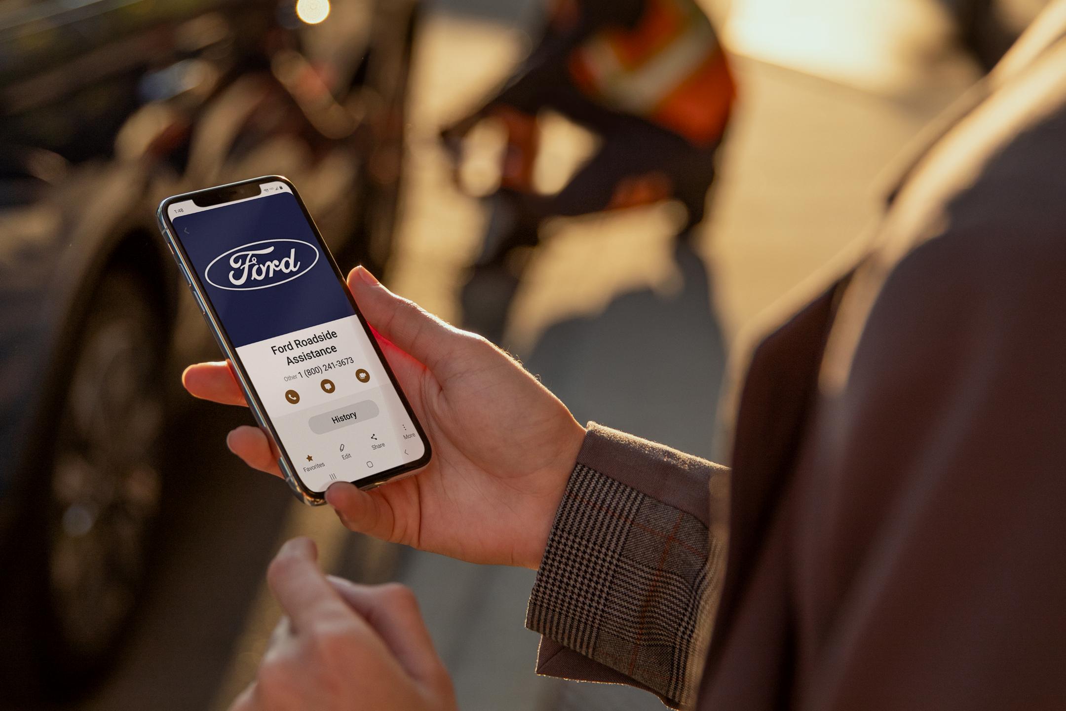Person holding a smartphone displaying a Ford Roadside Assistance screen.