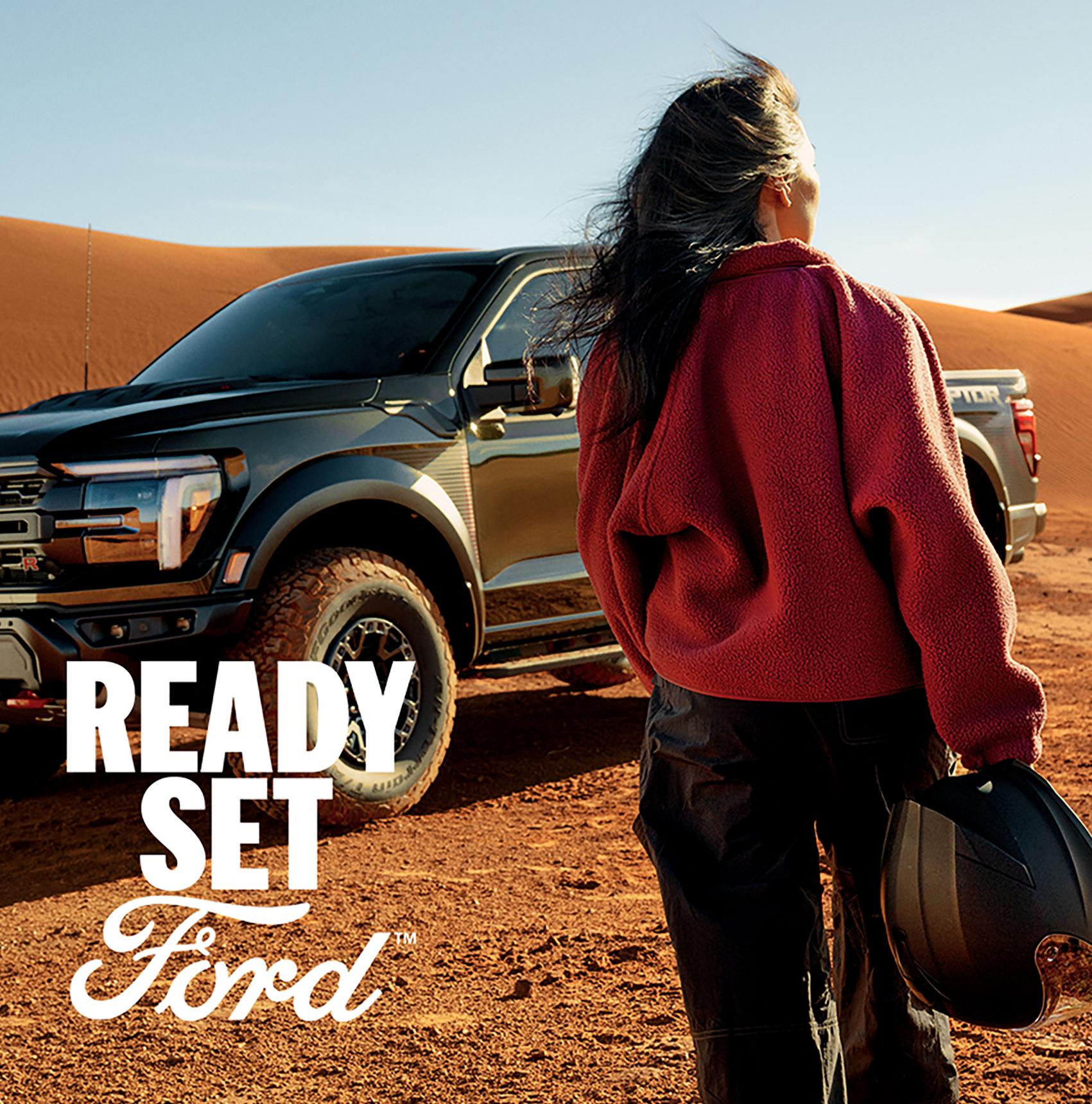 A woman, back turned, holds an off-road helmet while standing in the desert in front of a dark grey Ford F-150 Raptor. White "Ford" text is in the left corner.