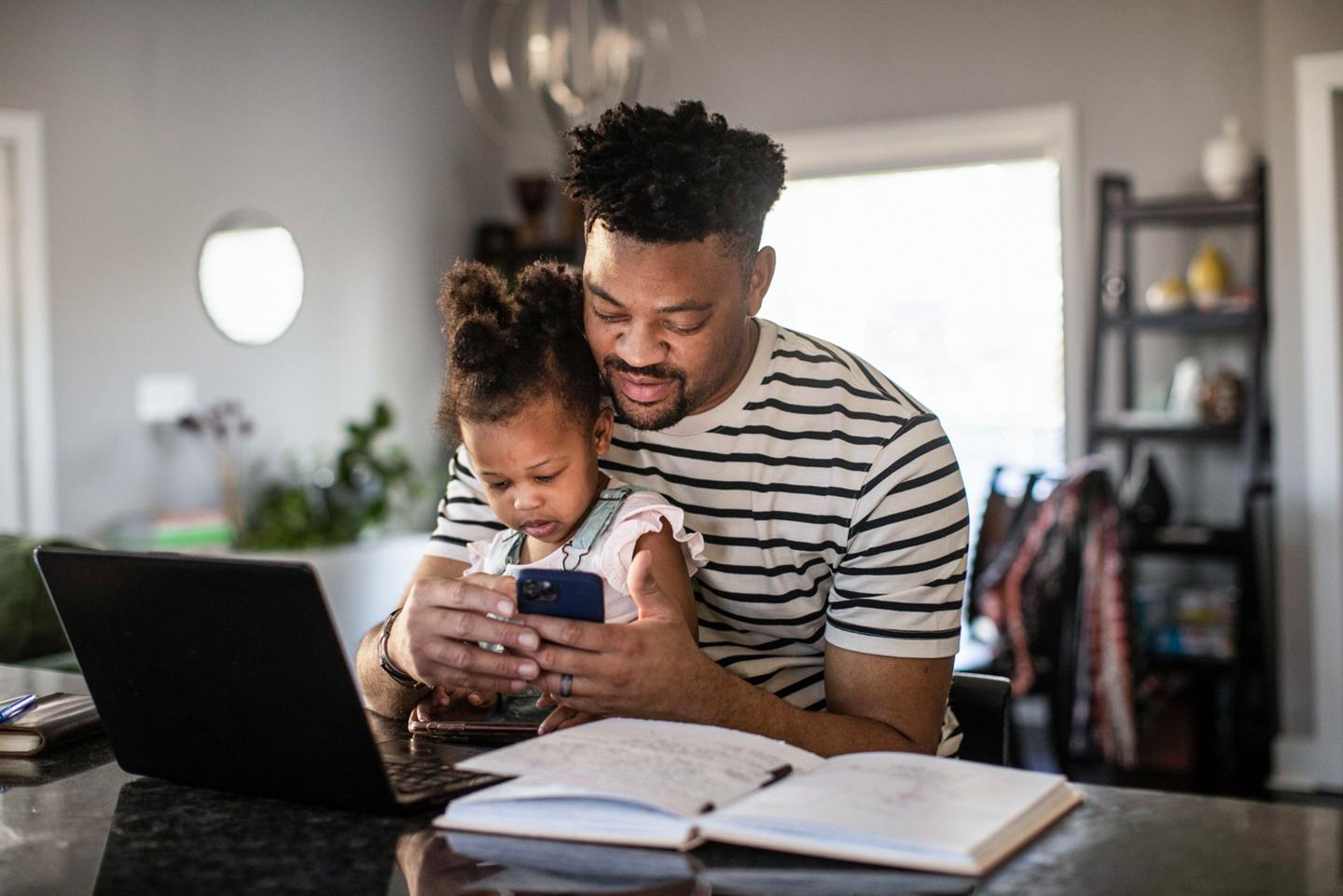 Man using his smartphone, while his daughter sits on his lap and looks on interestedly