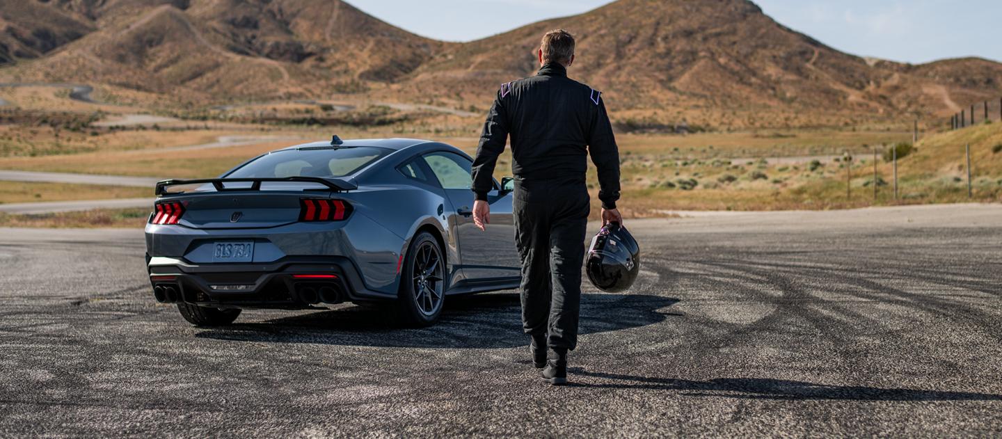 Person holding a racing helmet walks toward a Mustang Dark Horse parked on a track.