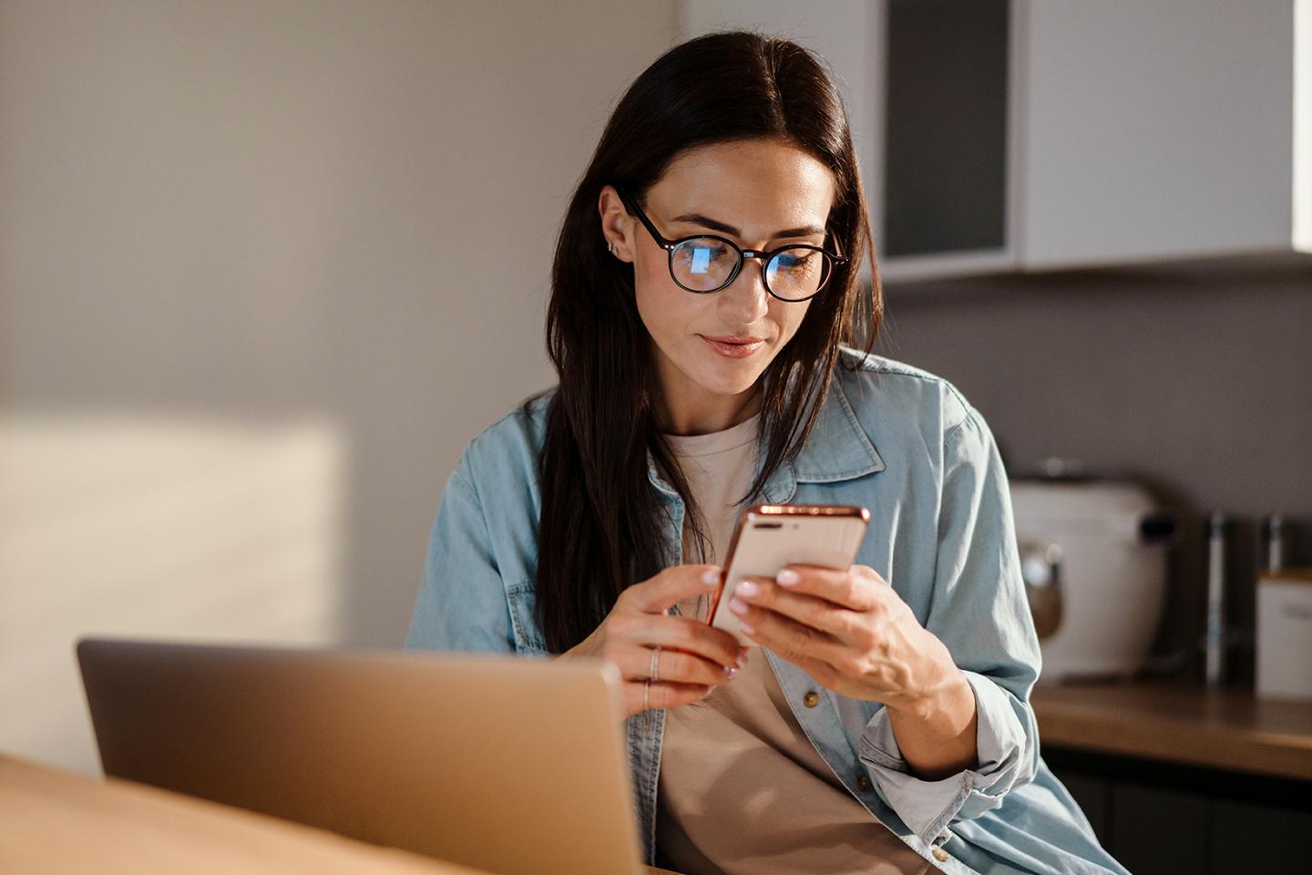 Person sits in front of a laptop while looking at their phone.