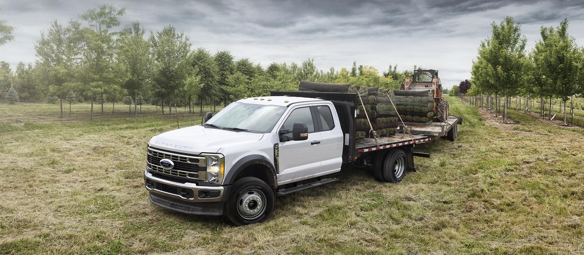 A White 2026 Ford Super Duty® Chassis Cab hauling something and towing a trailer with a wheeled skid steer
