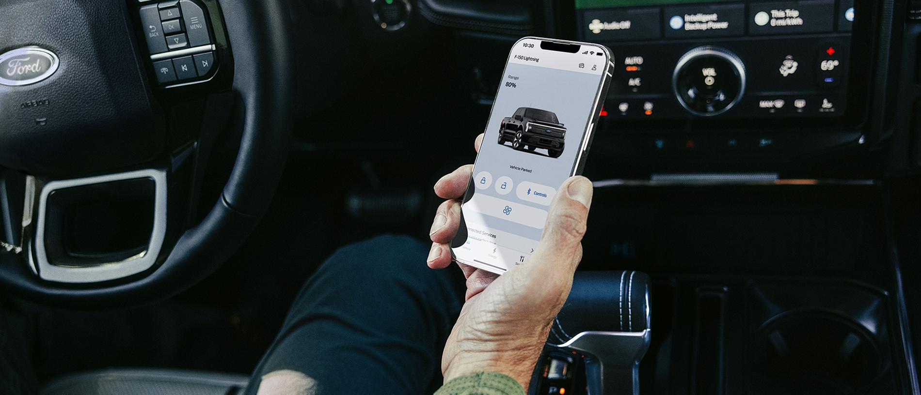 The hand of a driver seated in a 2025 Ford F-150® Lightning® pickup, holding a smartphone showing the FordPass® app