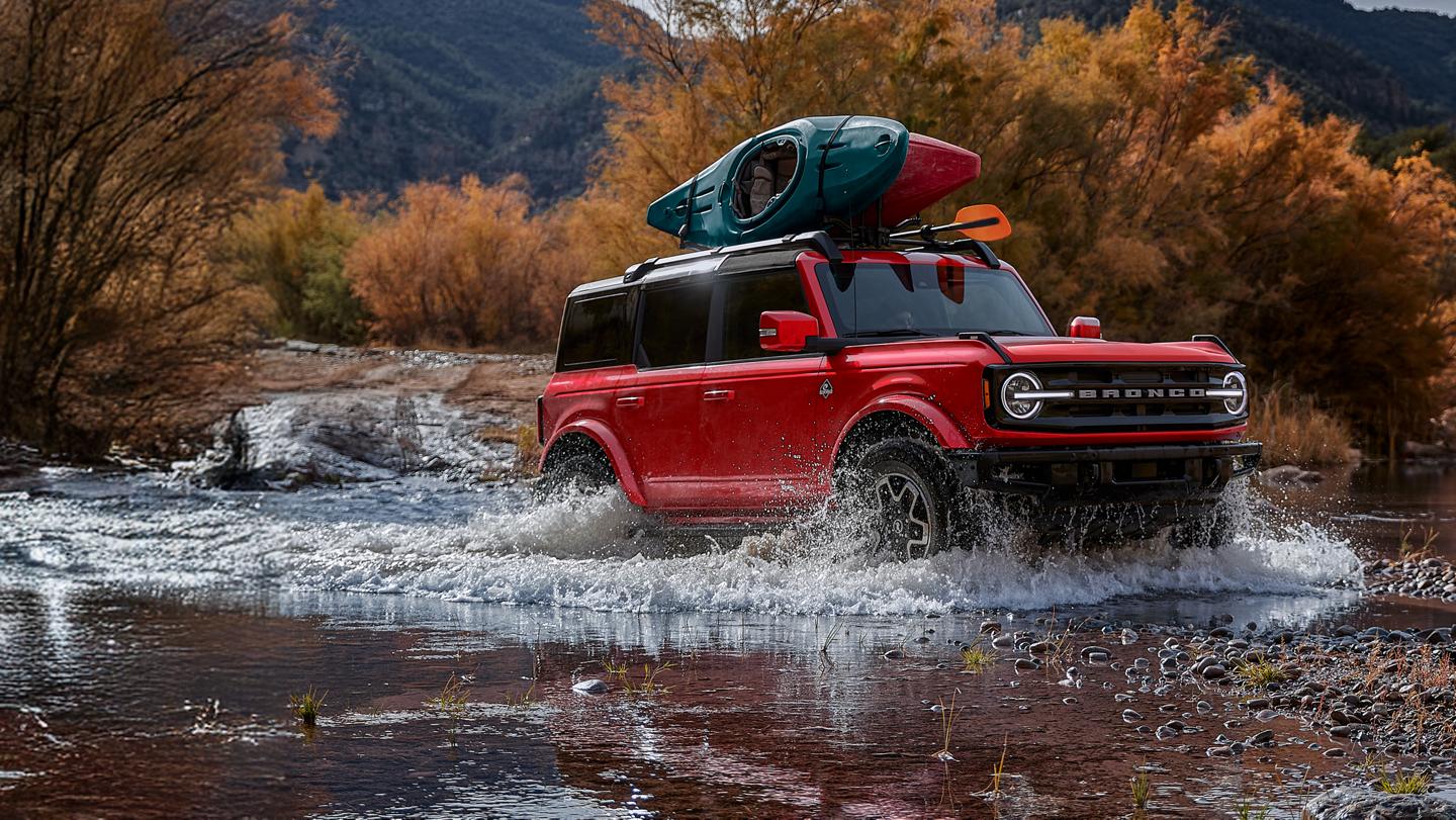Bronco with roof-mounted canoe driving through water.