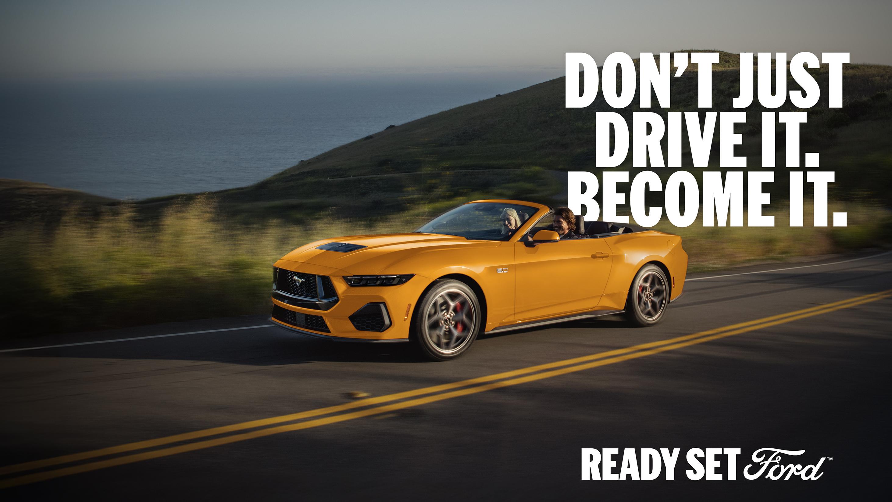 Two people in a 2026 Ford Mustang® GT Convertible in Orange Fury Metallic Tri-coat being driven on a coastal highway