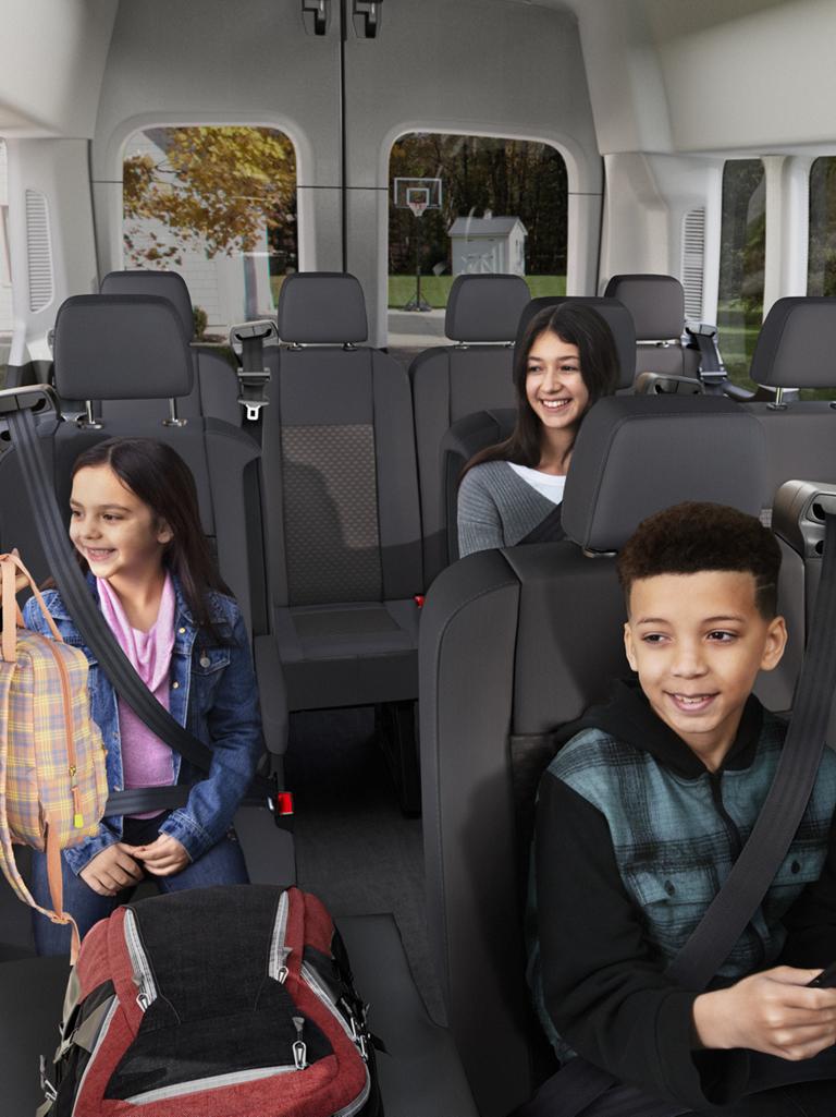 An adult woman, a young girl, and a young boy sit smiling in a multi-row interior of a 2027 Ford Transit® Passenger Van