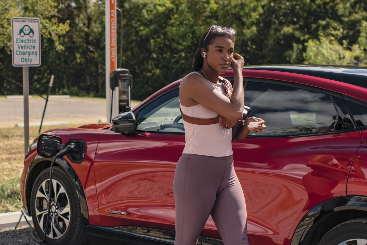 A woman walks away from a red Mustang Mach-E that is plugged into a public charging station.