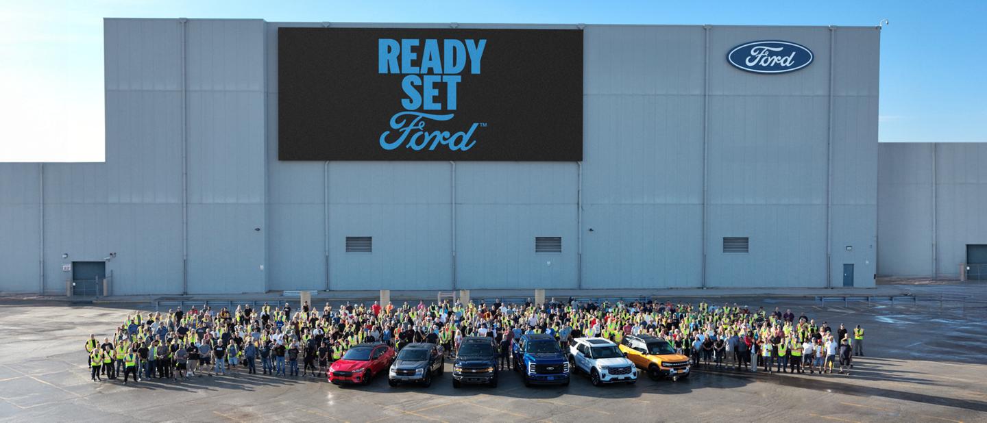 Wide shot of six parked Ford vehicles in front of a large group of Ford Employees at a dealership.