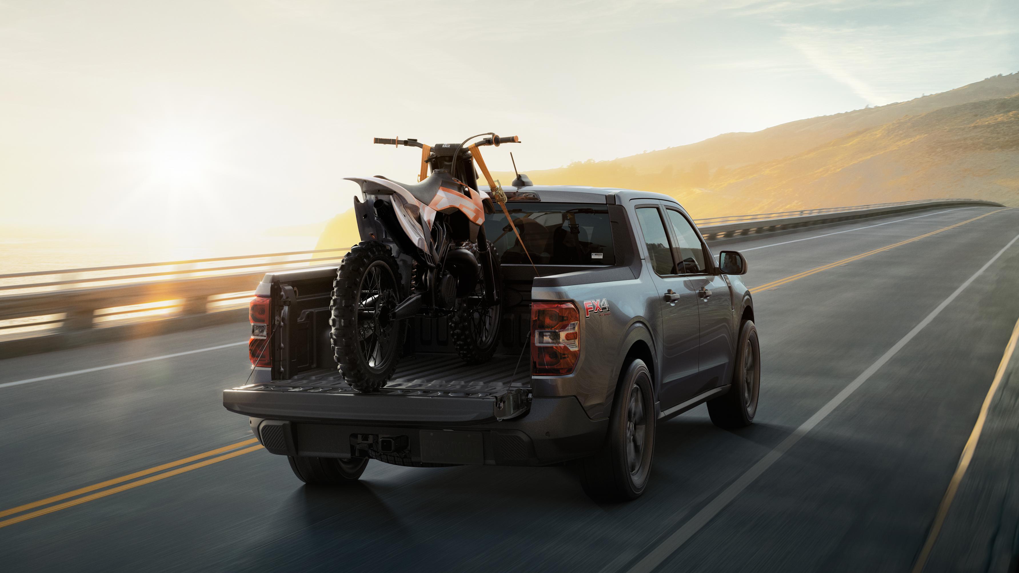 2026 Ford Maverick® pickup with a dirt bike secured in the bed being driven on a highway at sunrise