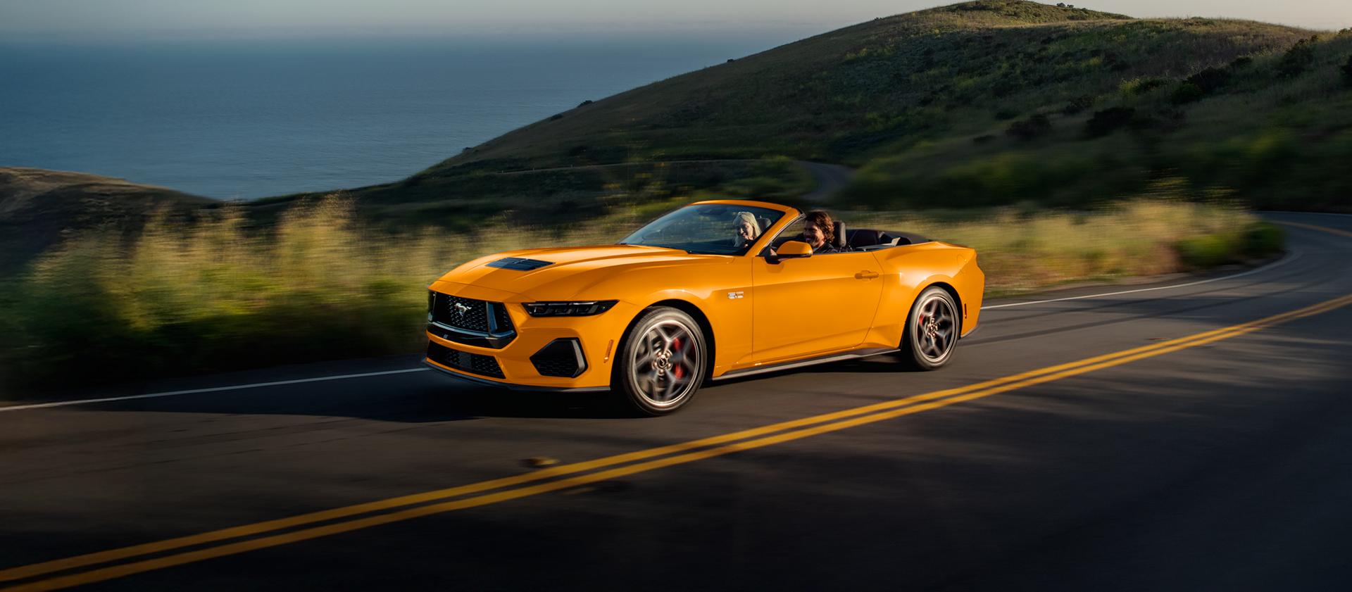 Two people in a 2026 Ford Mustang® GT convertible in Orange Fury Metallic Tri-coat being driven on a coastal highway