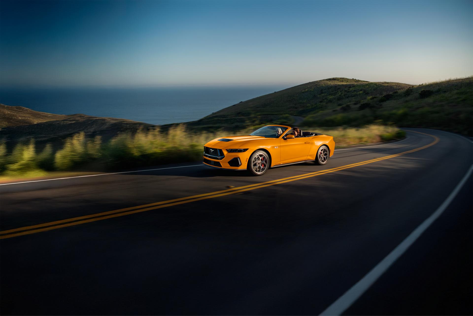 Two people in a 2026 Ford Mustang® GT convertible in Orange Fury Metallic Tri-coat being driven on a coastal highway