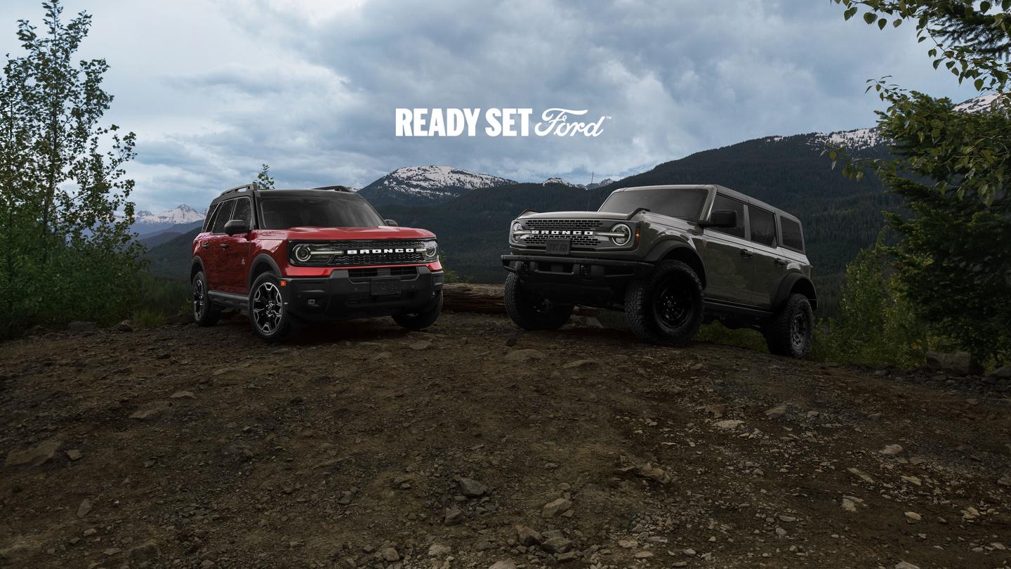 A red Ford Bronco Sport on the left and a grey four-door Bronco on the right, parked on a dirt hill with mountains in the background and the text 'READY SET Ford' above them.