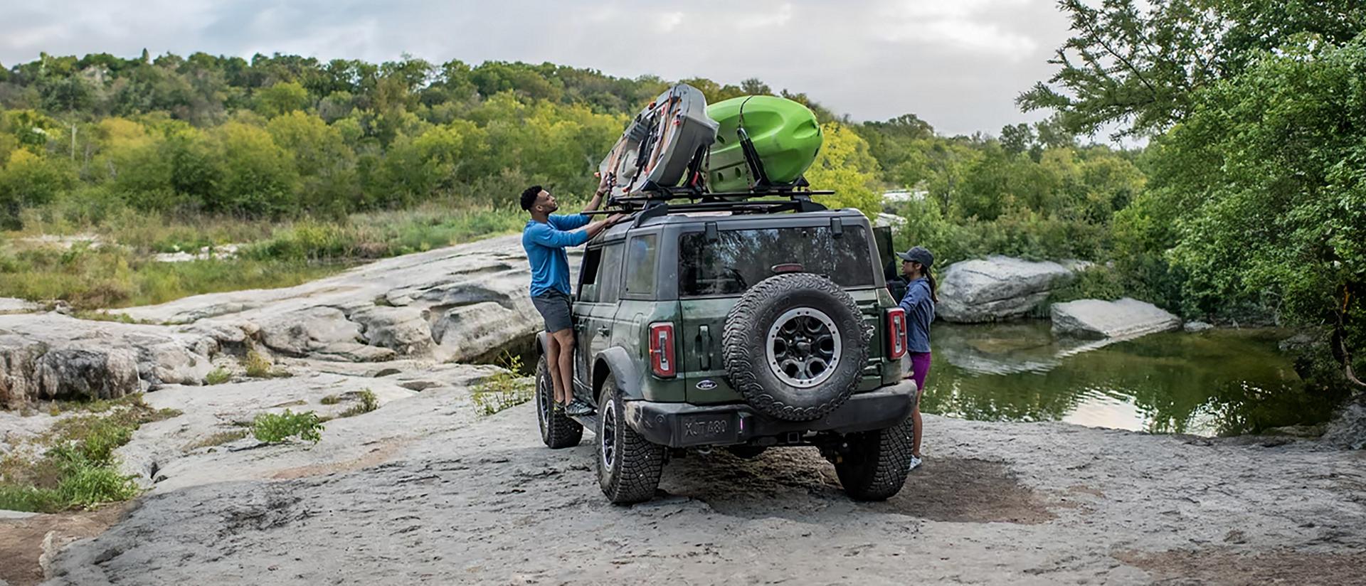 A man unloading an inflatable raft from the roof of a 2025 Ford Bronco® SUV parked on rocks near a stream