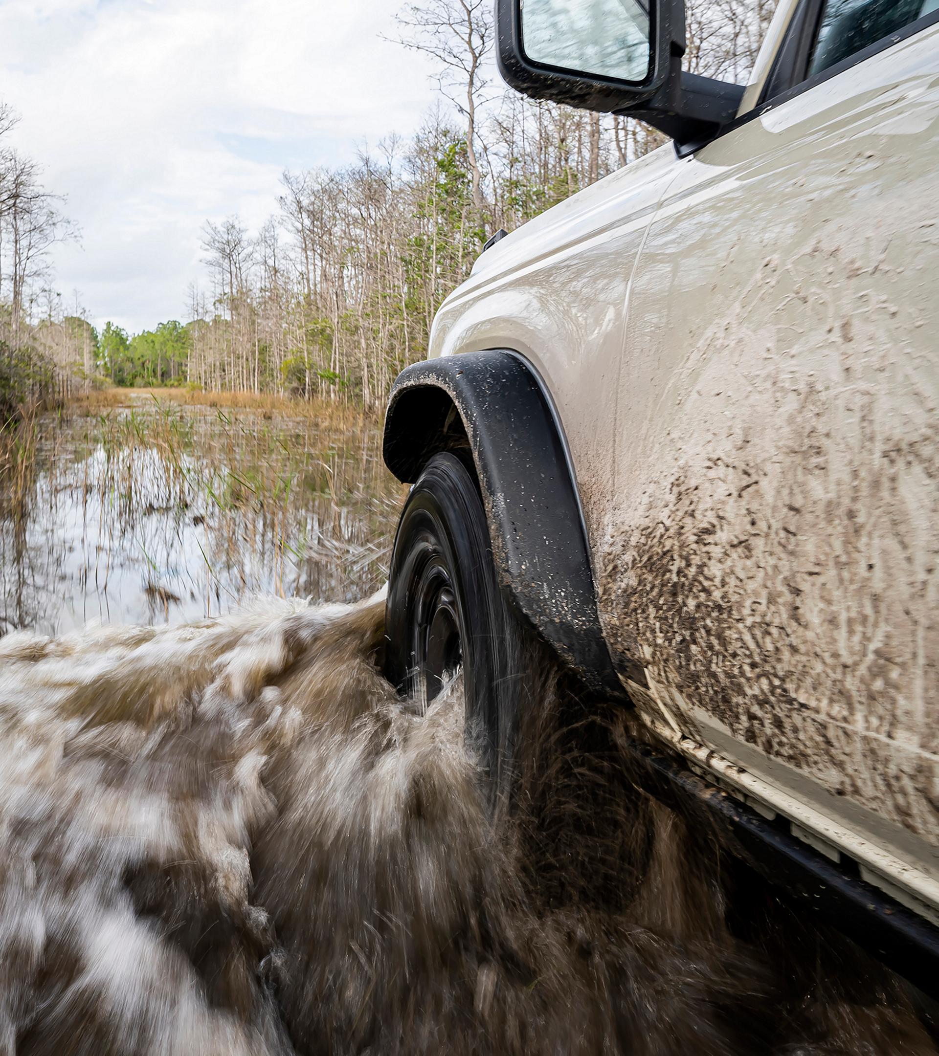 A close-up of the front driver's-side wheel and fender of a 2026 Ford Bronco® SUV as it's driven through a river