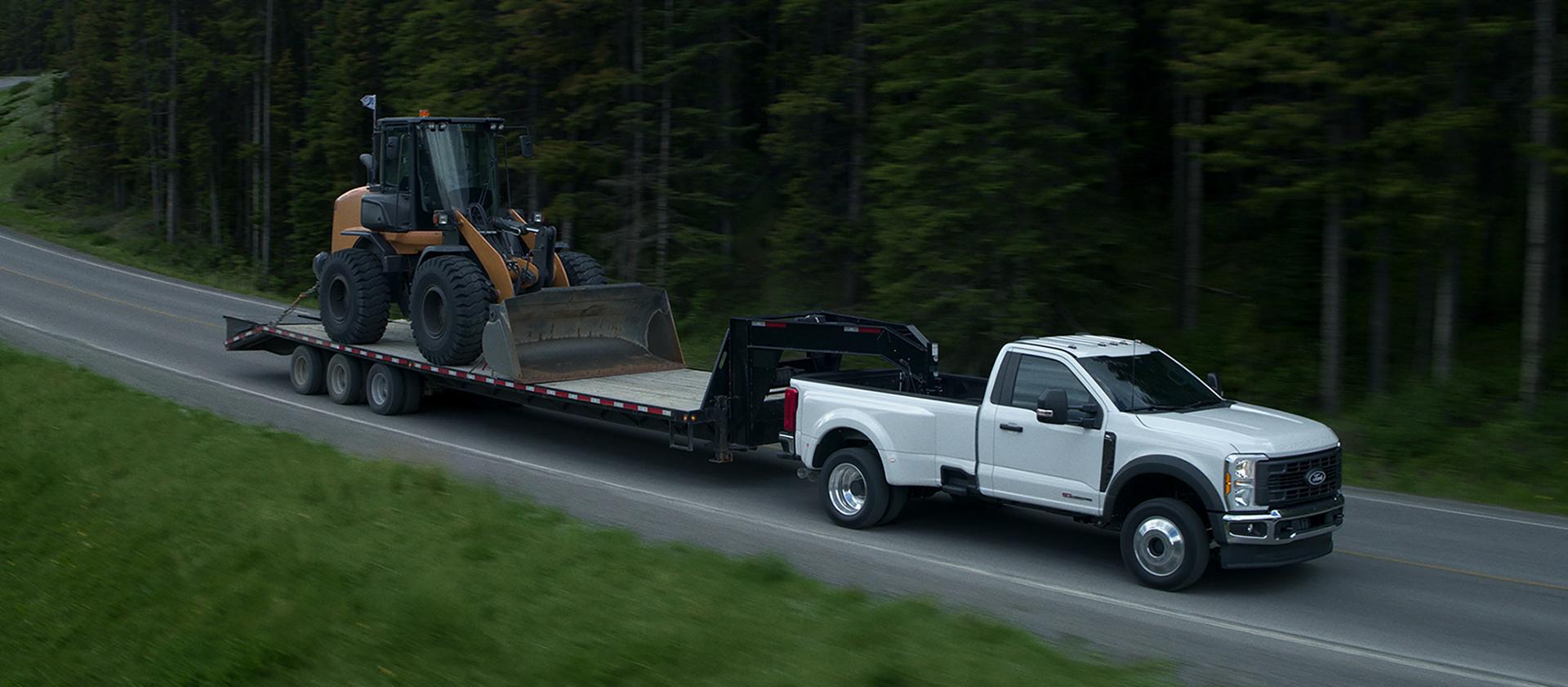 2027 Ford Super Duty® F-450® XL model towing a flatbed trailer carrying a compact wheel loader on a paved mountain road