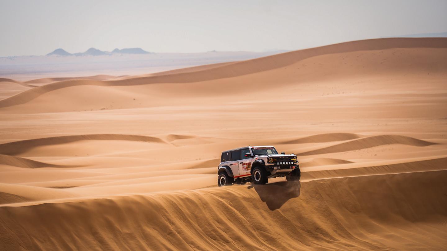 A Ford Bronco drives over dunes in a desert.