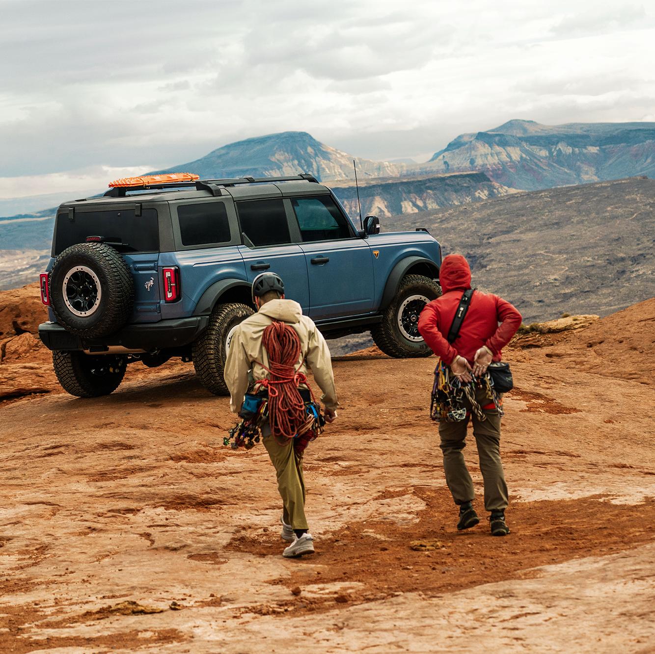 Two male climbers walking towards a Ford Bronco parked on rocky terrain.