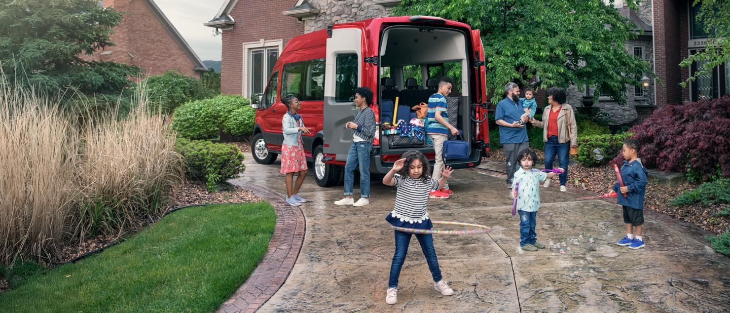 Two parents and a group of children stand behind a red Ford Transit Passenger Van.