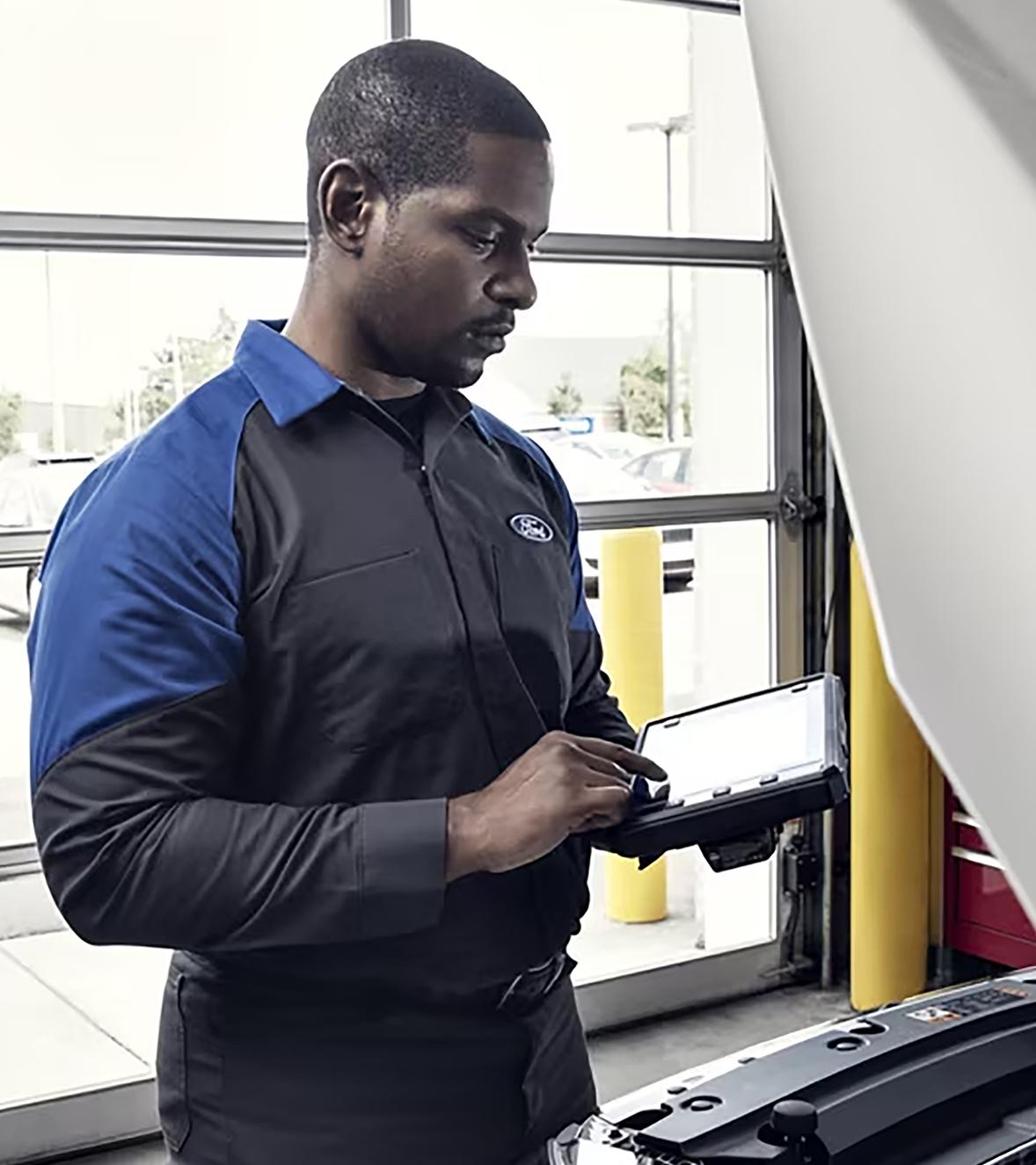 A Ford Service technician makes an entry on a tablet while looking under the hood of a Ford vehicle in the service bay.