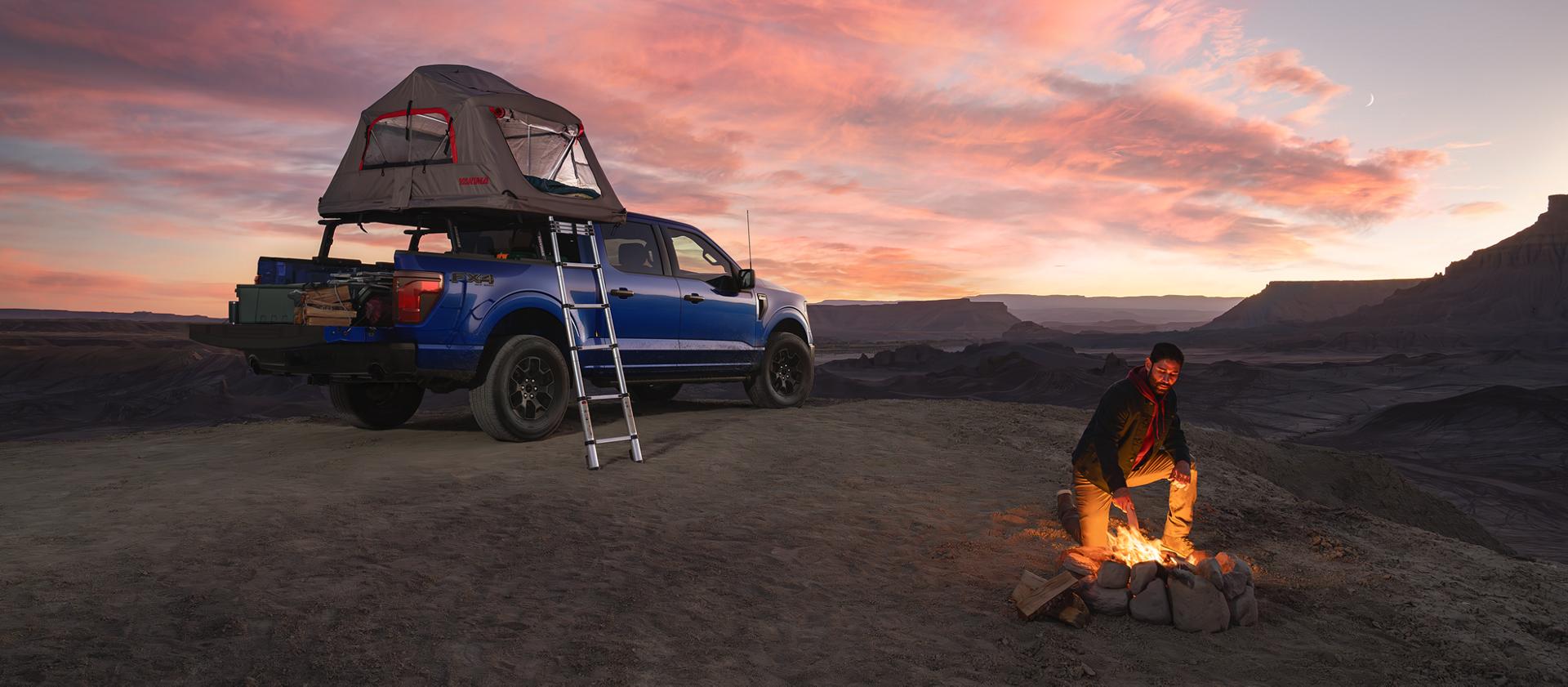 Man starting a camp fire near a parked 2026 Ford F-150® STX® FX4 model with a Ford Accessories tent at dusk