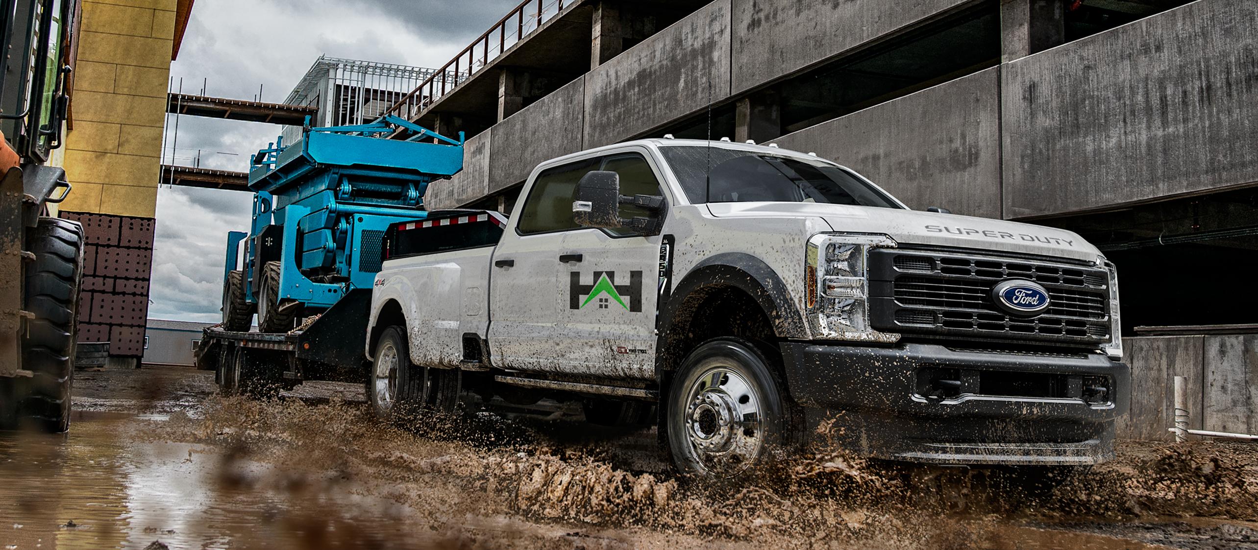 2026 Ford Super Duty® F-450® XL model pulling a flatbed trailer with a large blue tractor on it through mud and water