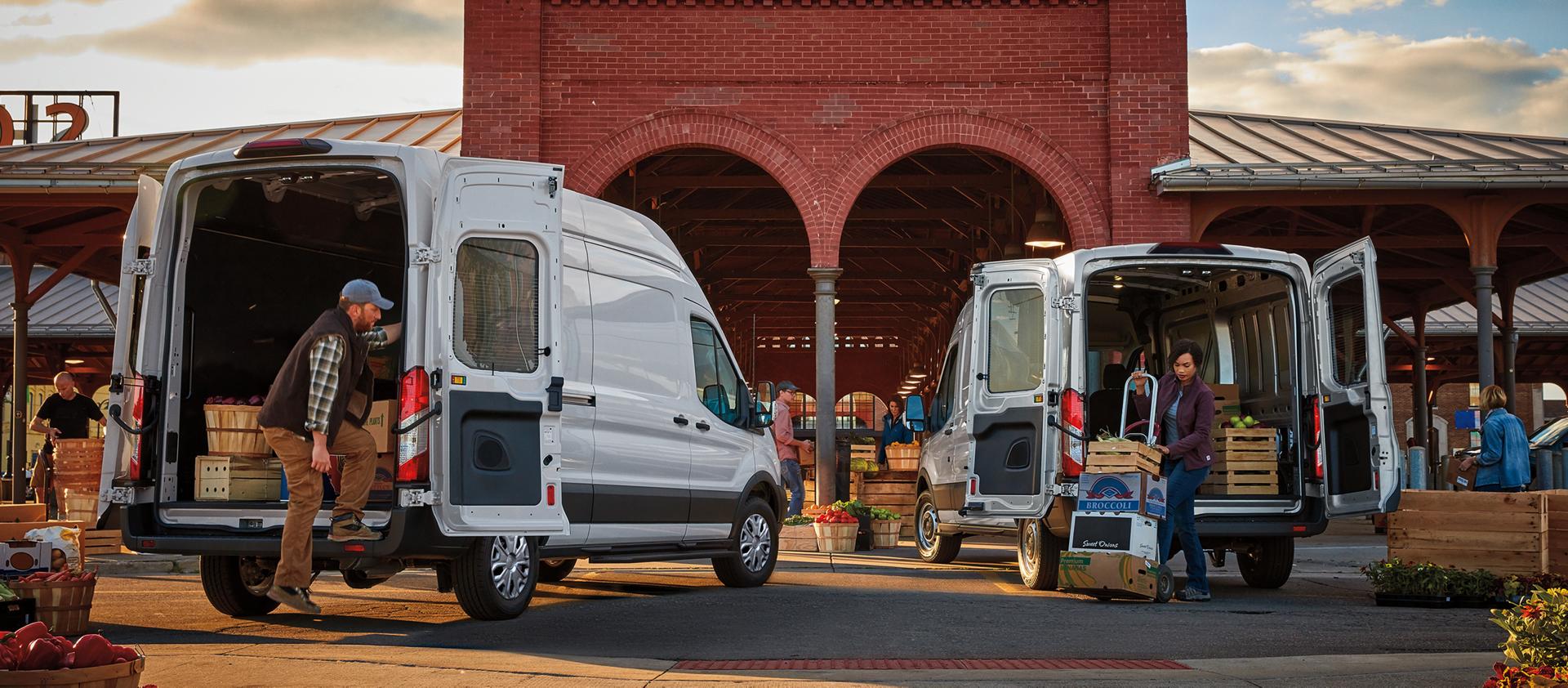 Two white Ford Transit® Vans with rear open and people unloading produce at a market