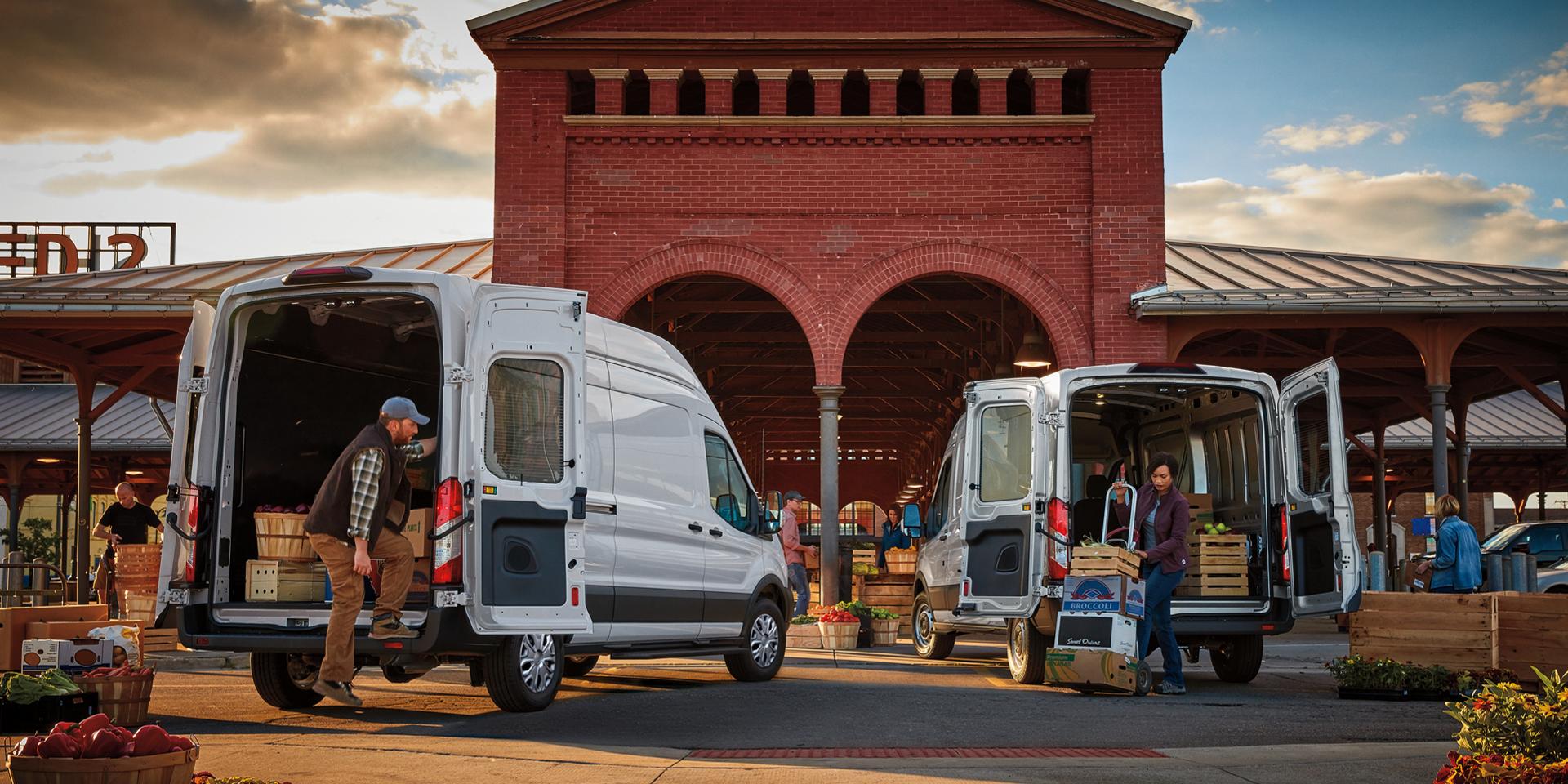 Two white 2027 Ford Transit® Vans with rear open and people unloading produce at a market