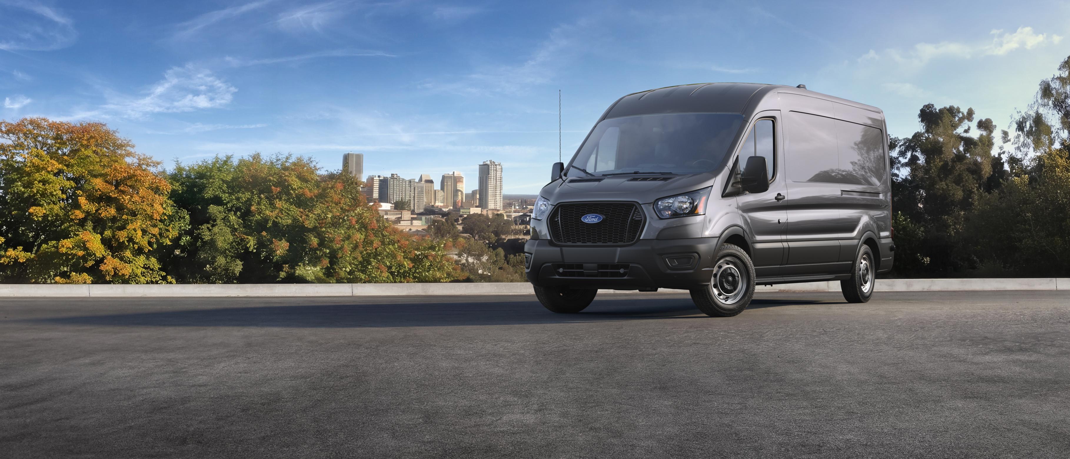 A grey 2027 Ford Transit® Cargo Van on a road with a city skyline and trees in the background