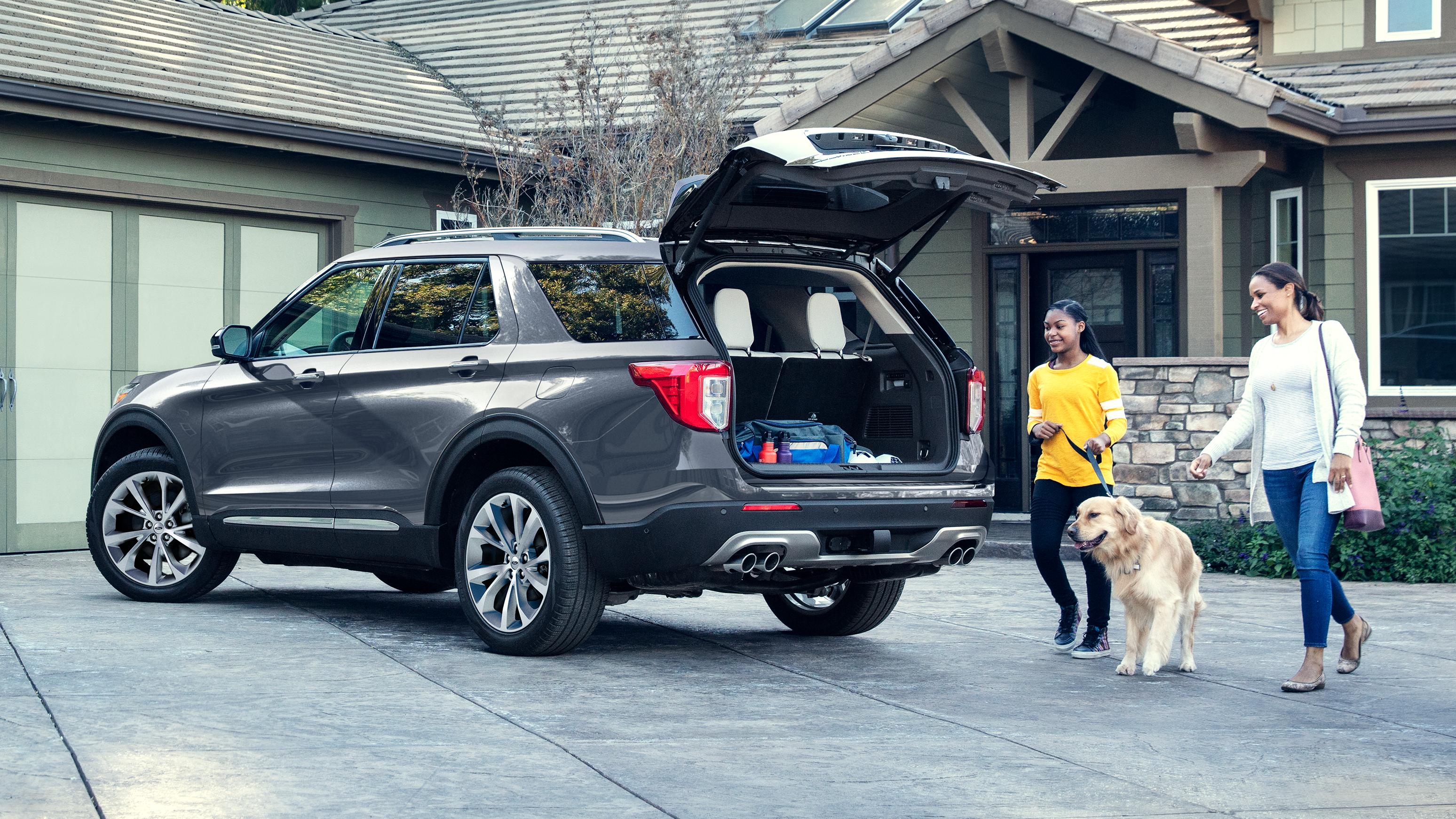 Two smiling people walk their dog over to the open liftgate of a Ford Explorer® SUV parked in a driveway.