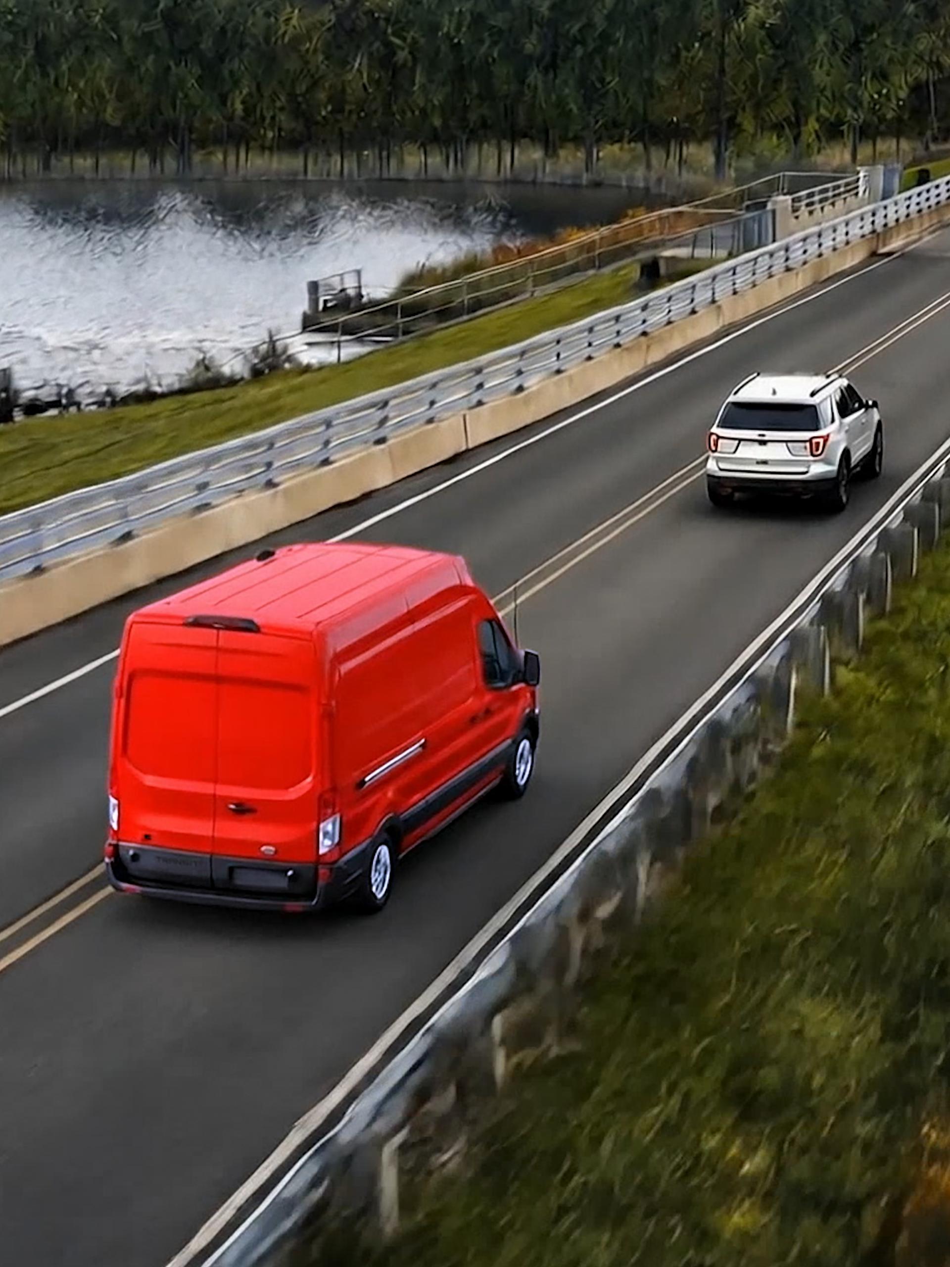 A Red 2027 Ford Transit® Van driving on a road alongside a river, with a white SUV ahead