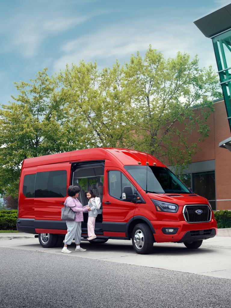 A red 2027 Ford Transit® Passenger Van parked outside a modern building with a family nearby