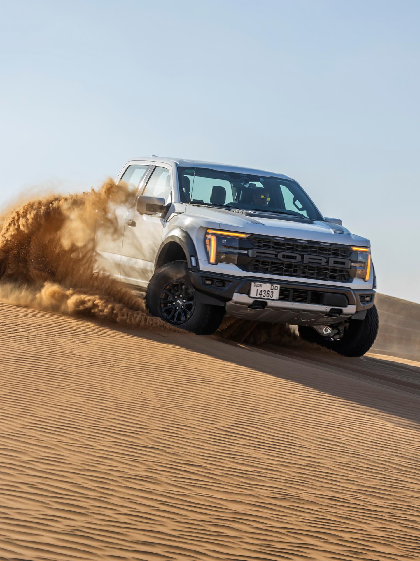 A dark blue Ford F-150 Raptor driving through sand and dust on a desert dune.